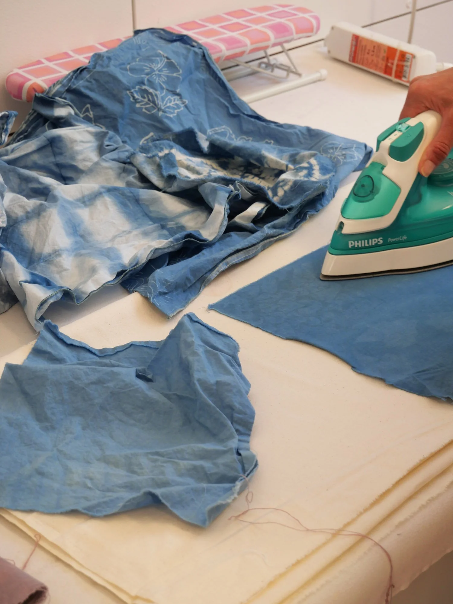 Plans, scraps of blue fabric, and an iron on a table, with an ironing board in the background.