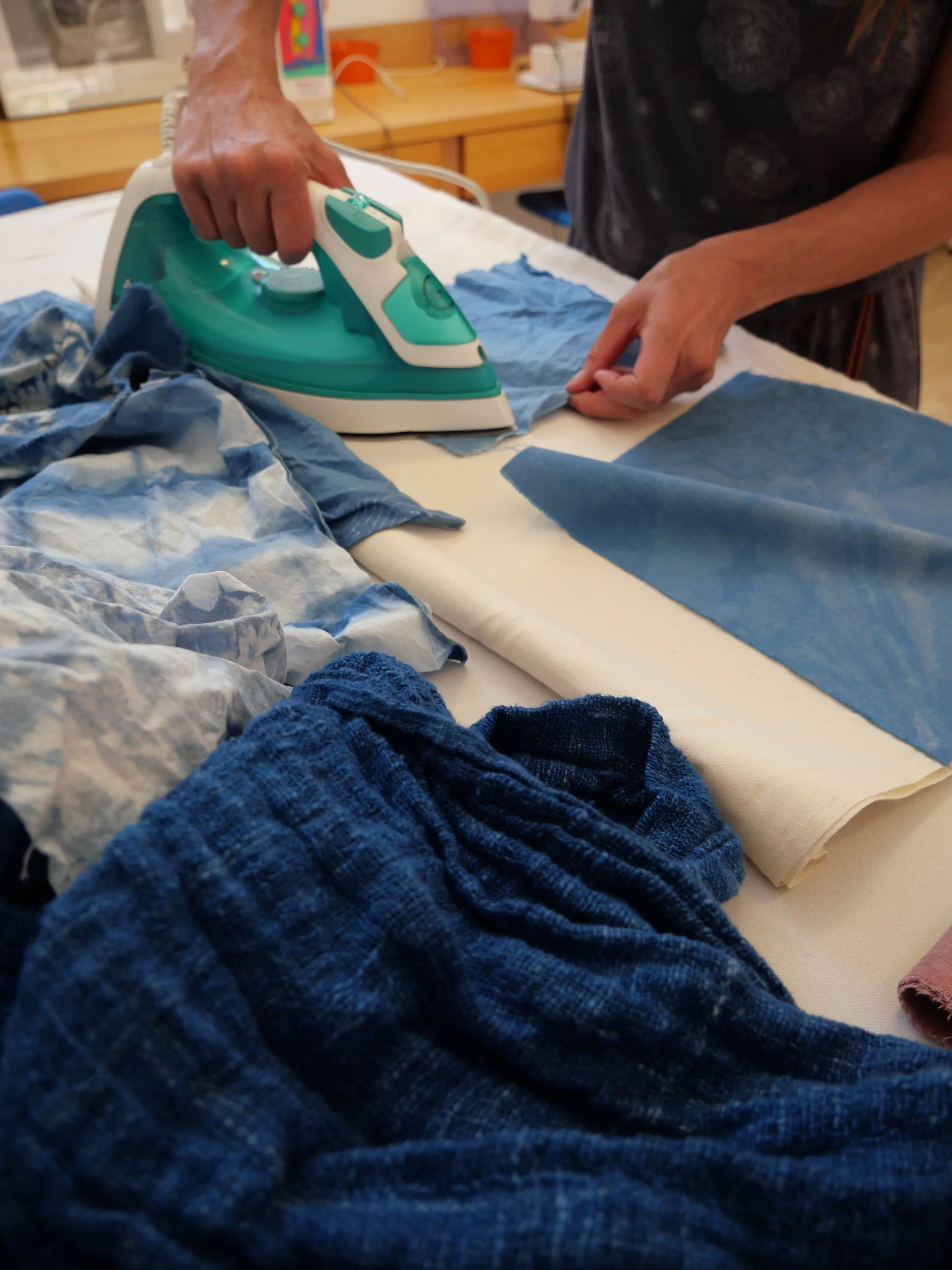 Une personne repasse des vêtements bleus avec un fer à repasser sur une table. Autour, on voit des tissus et des vêtements en désordre.