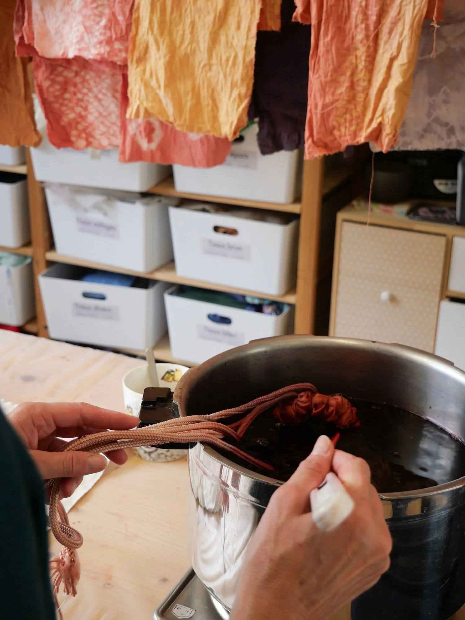 Une personne manipule une corde de coton pour teindre du tissu dans un bain de teinture chaud dans une cuisine ou un atelier, avec plusieurs vêtements colorés suspendus pour sécher en arrière-plan.