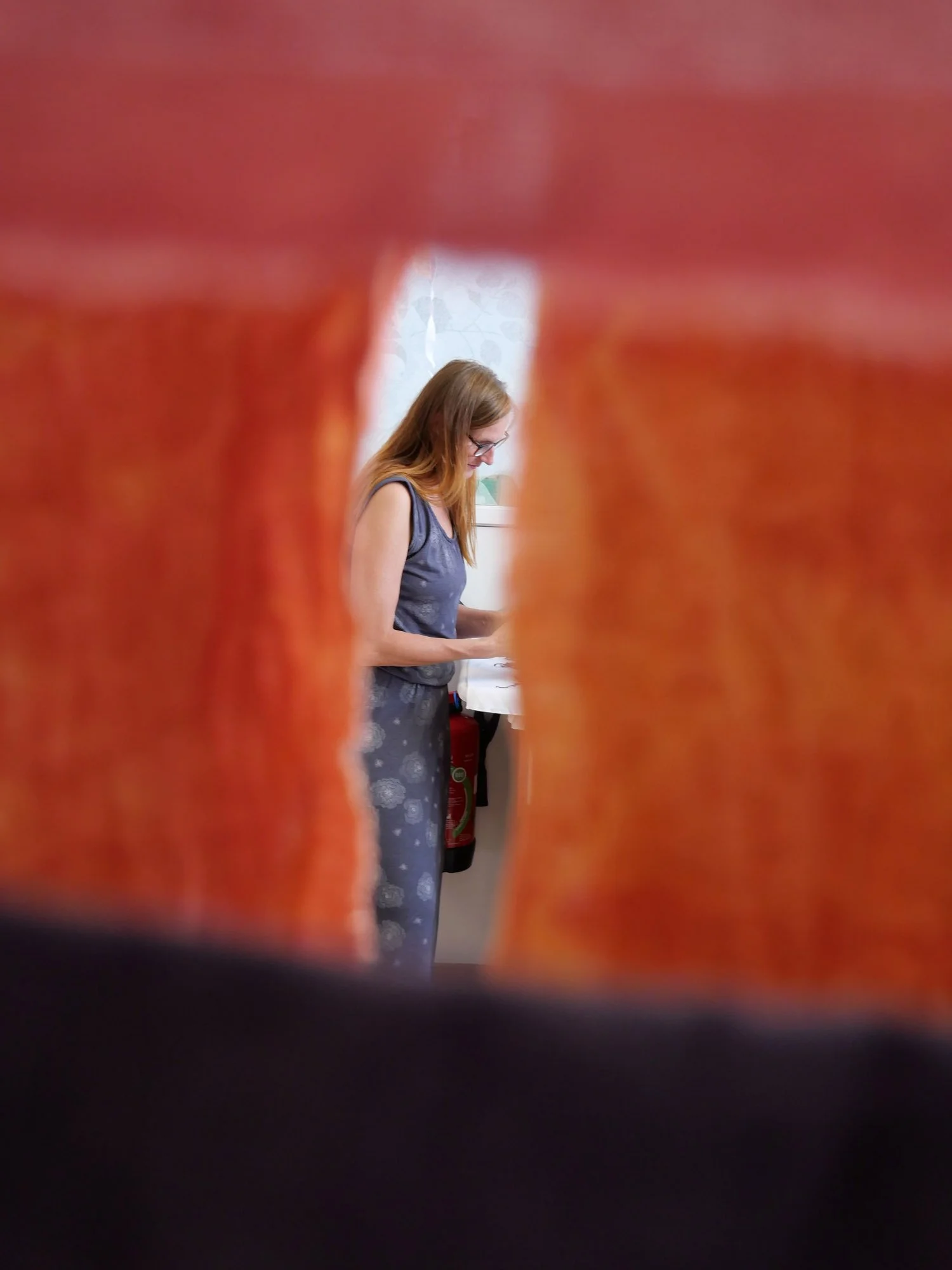 Une femme aux cheveux roux portant une robe grise, vue à travers un trou dans un vêtement orange, en train de lire ou d'écrire quelque chose, avec un évier en arrière-plan.