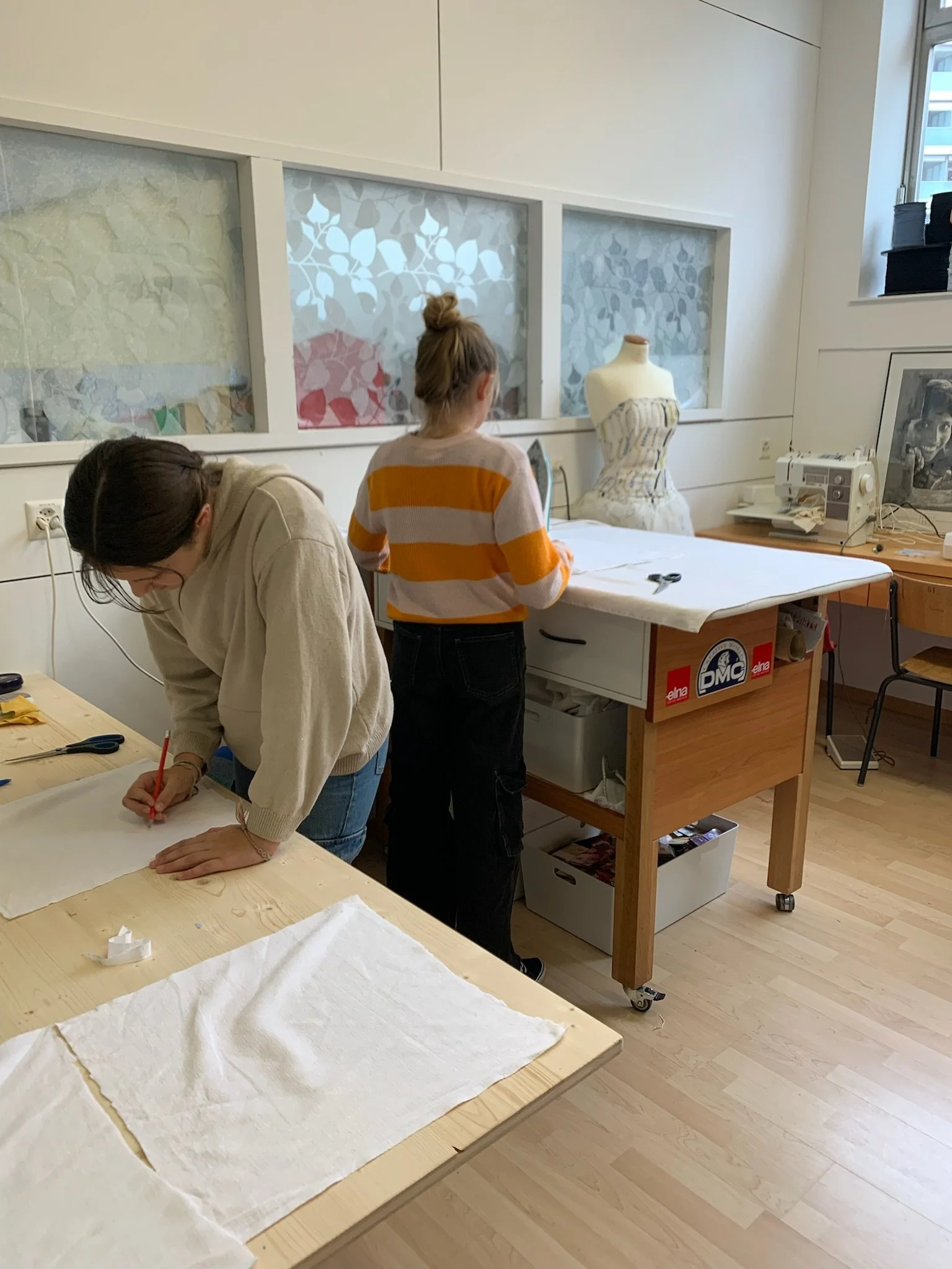 Deux femmes travaillent dans un atelier de couture avec un mannequin habillé d'une robe et une table de couture équipée d'une machine à coudre et de ciseaux.