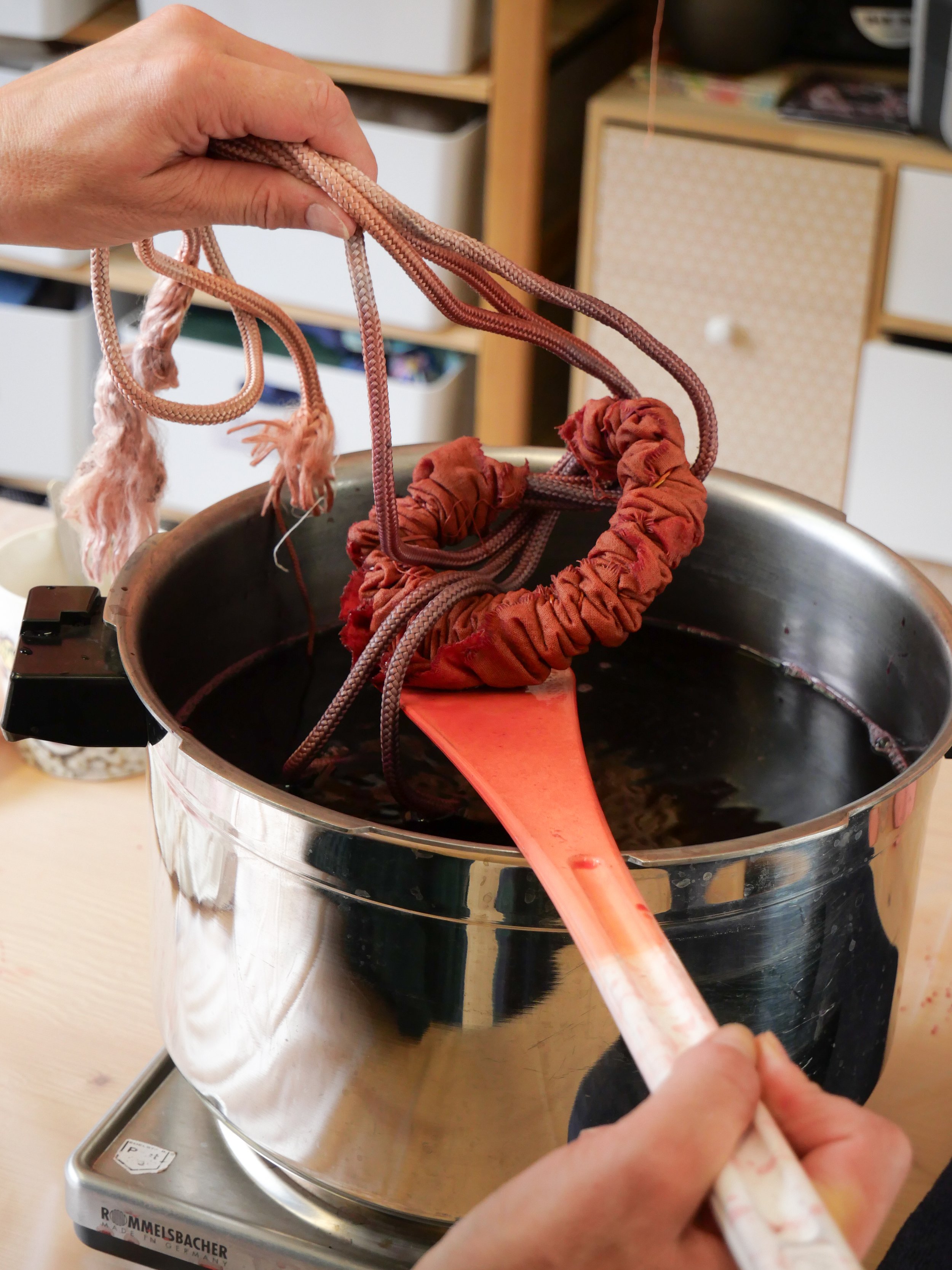 Une main utilise une spatule rouge pour manipuler des cordes en tissu suspendues dans un bain de teinture noire, dans un contexte de teinture textile.