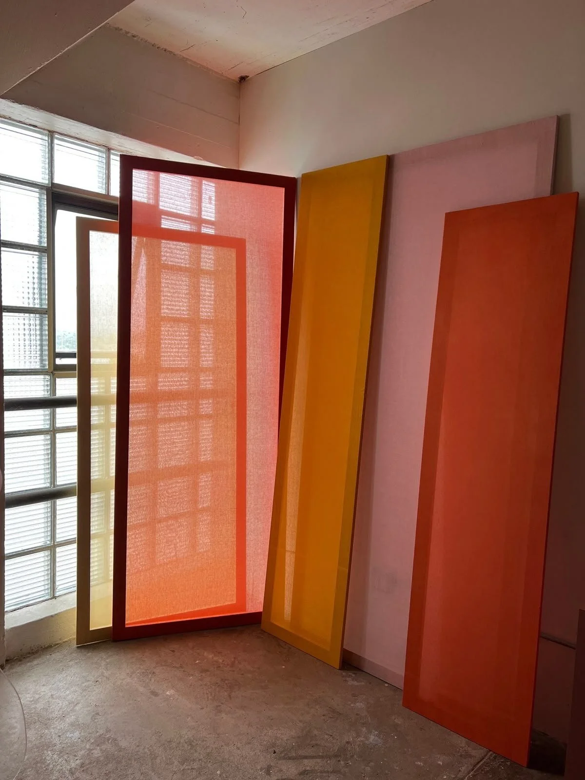 Plusieurs cadres colorés, en rose, orange, jaune, et rouge, posés contre un mur près d'une fenêtre en verre.