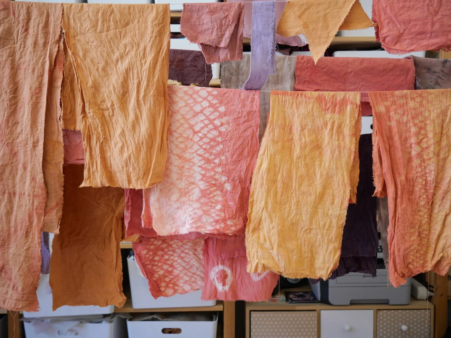 Des tissus colorés en teintes de pêche, orange et lavande suspendus pour sécher, dans une pièce avec des meubles en bois.