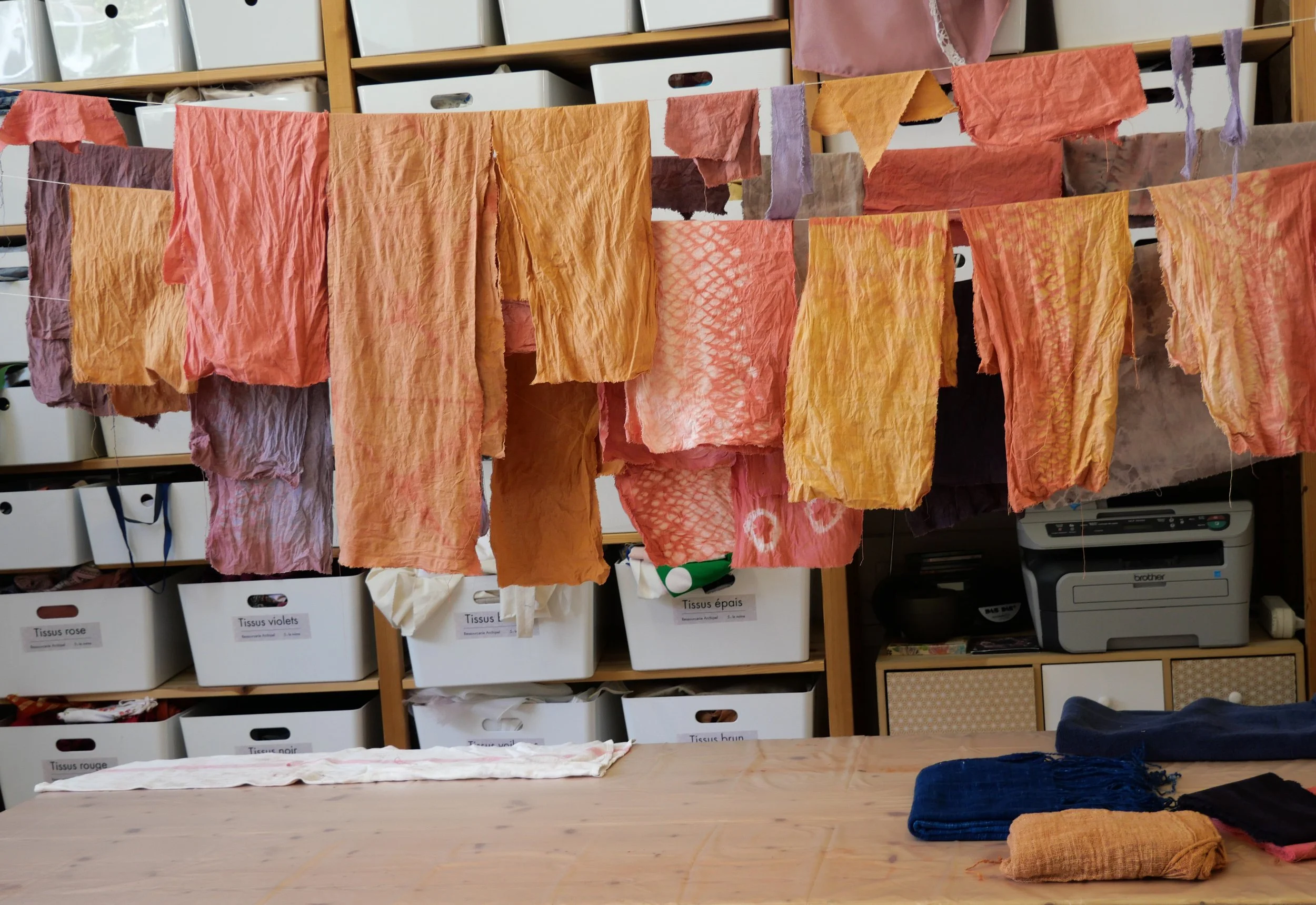 Tissus en coton de différentes couleurs suspendus à une corde, dans une pièce avec étagères de rangement et imprimante.