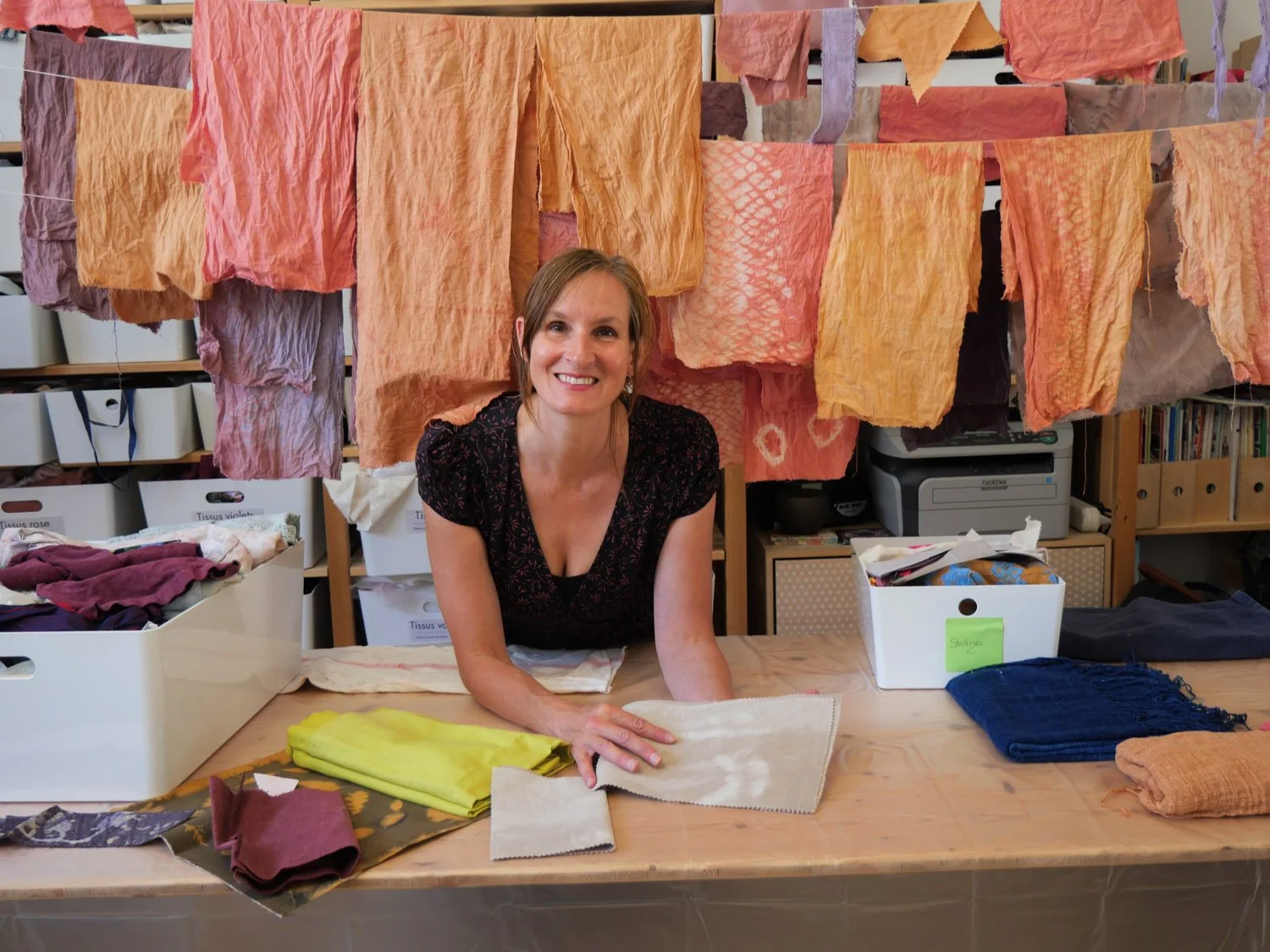 Une femme souriante présente à un atelier de teinture avec des tissus colorés suspendus en arrière-plan.
