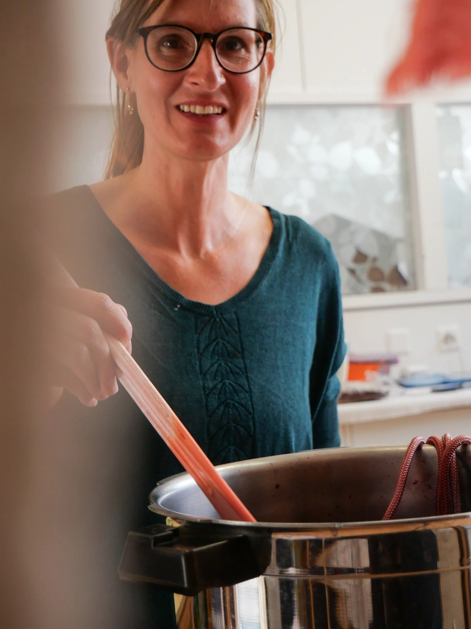 Une femme souriante portant des lunettes, utilisant une pince pour manipuler quelque chose dans une marmite en cuisine.