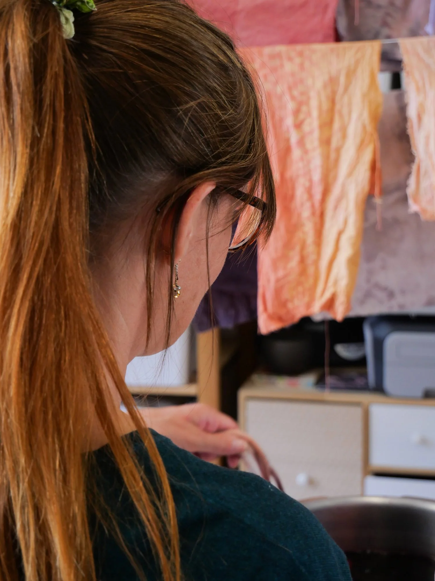 Une femme vue de dos avec des cheveux roux attachés, portant des lunettes et des boucles d'oreilles, travaille près d'un lavabo avec un linge suspendu en arrière-plan.