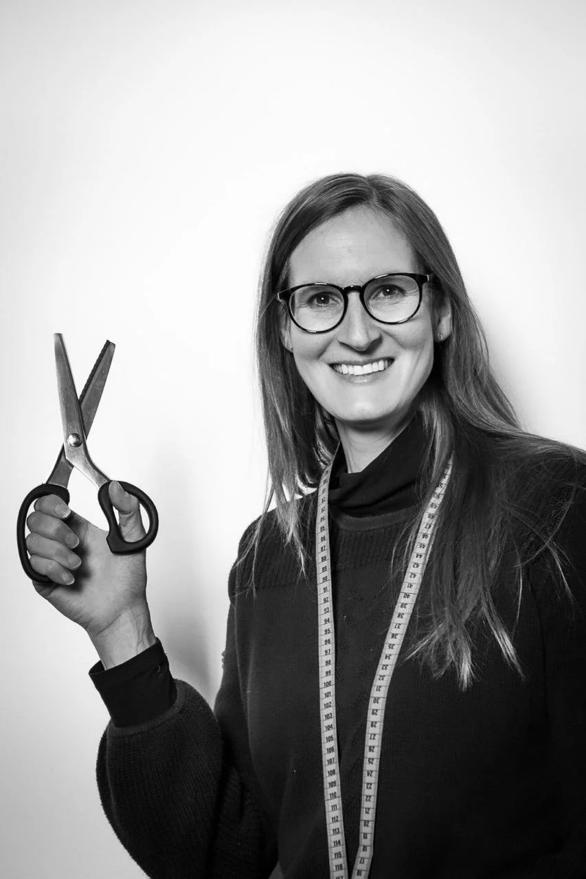 Une femme souriante portant des lunettes, une robe noire, avec un ruban à mesurer autour du cou, tenant une paire de ciseaux.