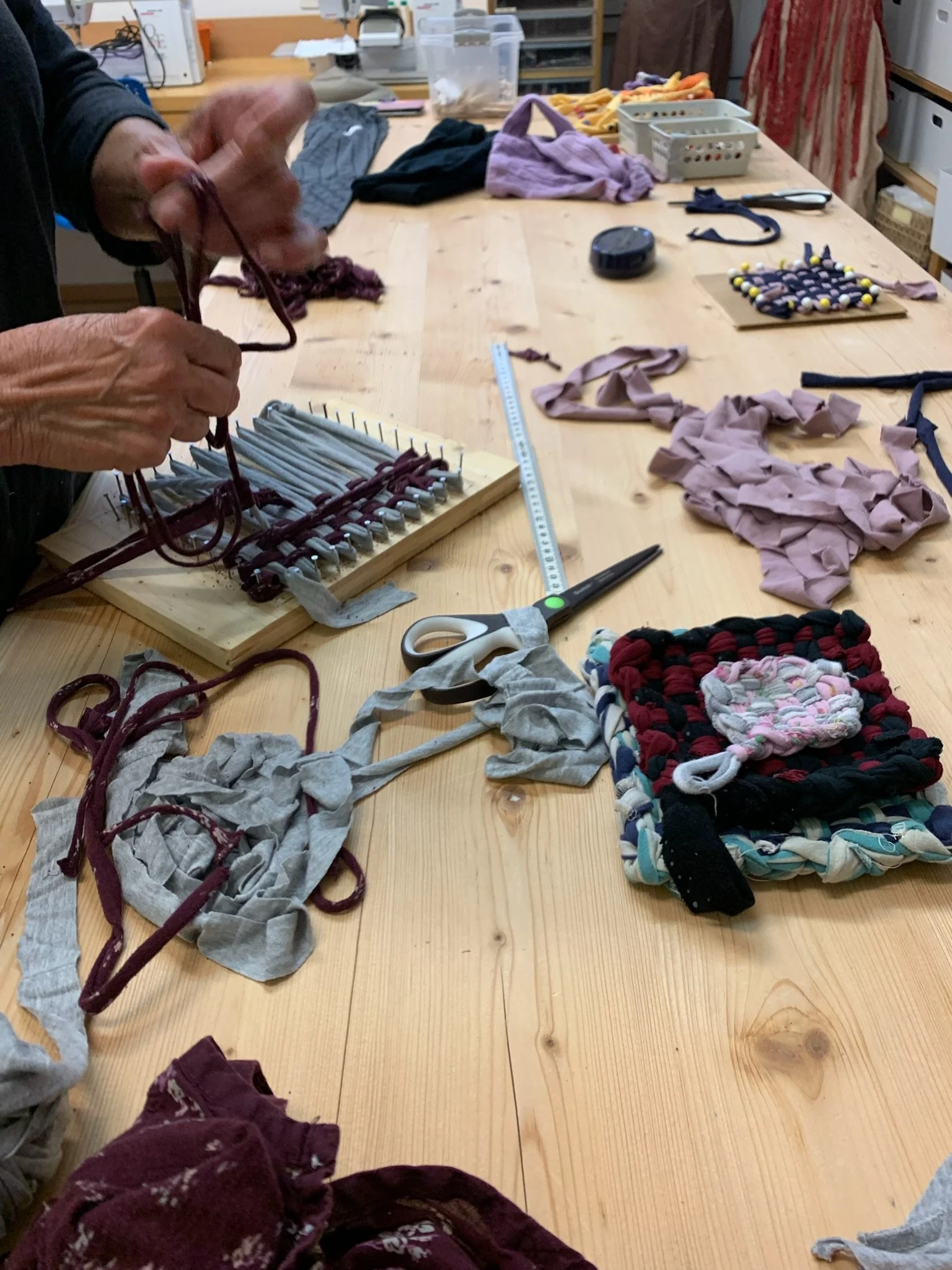 Une personne assemble des morceaux de tissu pour créer des objets textiles, entourée de divers tissus, rubans, ciseaux, règles, et autres outils sur une grande table en bois.