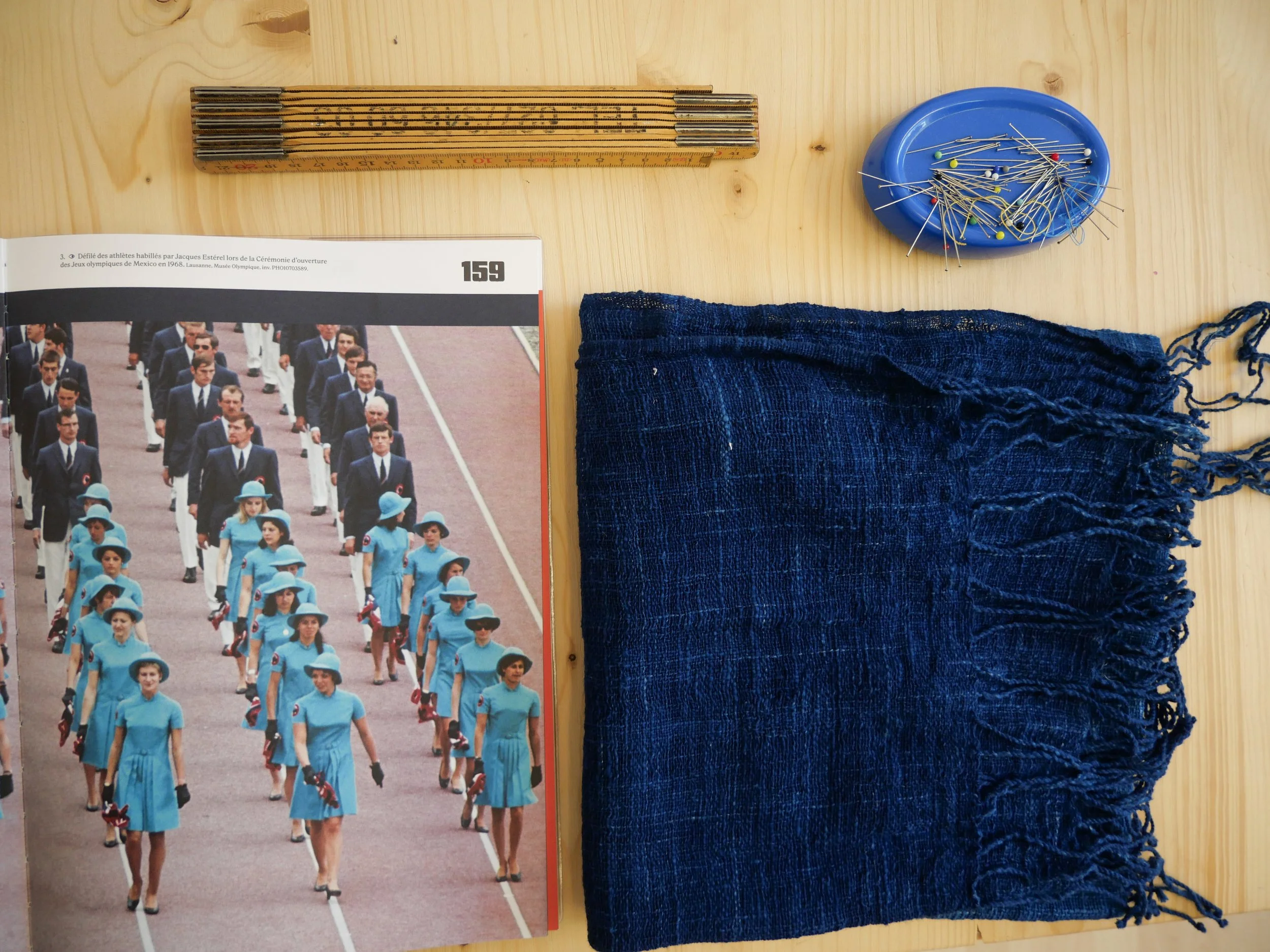 Une photo montrant une répartition d'objets sur une surface en bois clair, comprenant une règle en bois, une brochure ou photo d'une parade militaire avec des personnes en uniforme bleu, un sac en tissu bleu foncé, une poubelle à épingles avec plusie