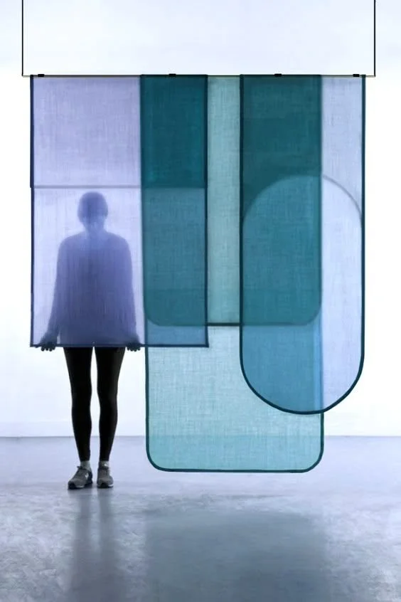 Une personne silhouette derrière des panneaux en tissu translucide suspendus, dans un espace blanc lumineux.