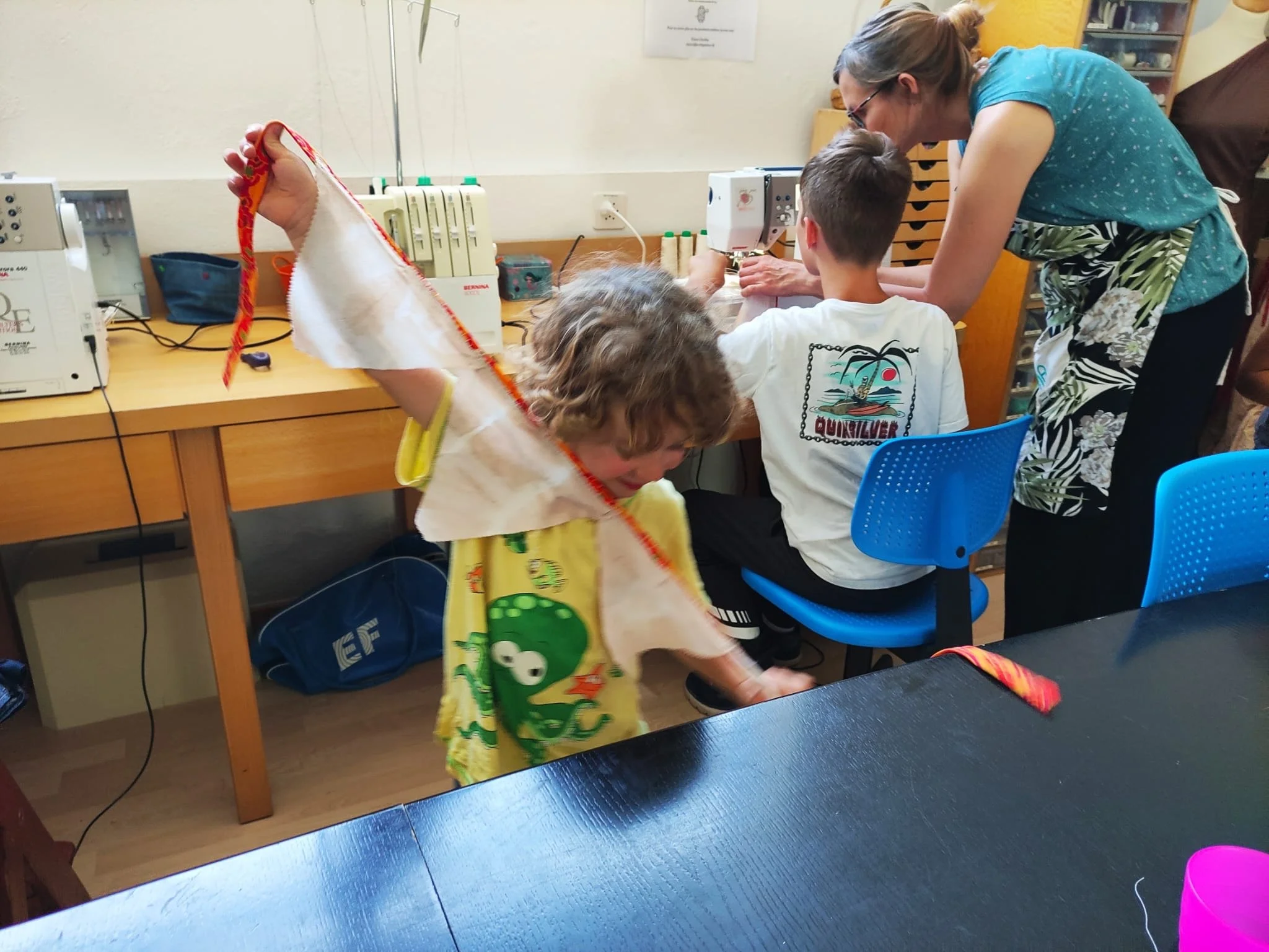 Une femme et deux enfants dans un atelier. La femme aide un enfant assis à la machine à coudre. Un autre enfant, portant une cape en tissu blanc avec des bordures rouges, joue près du tableau.