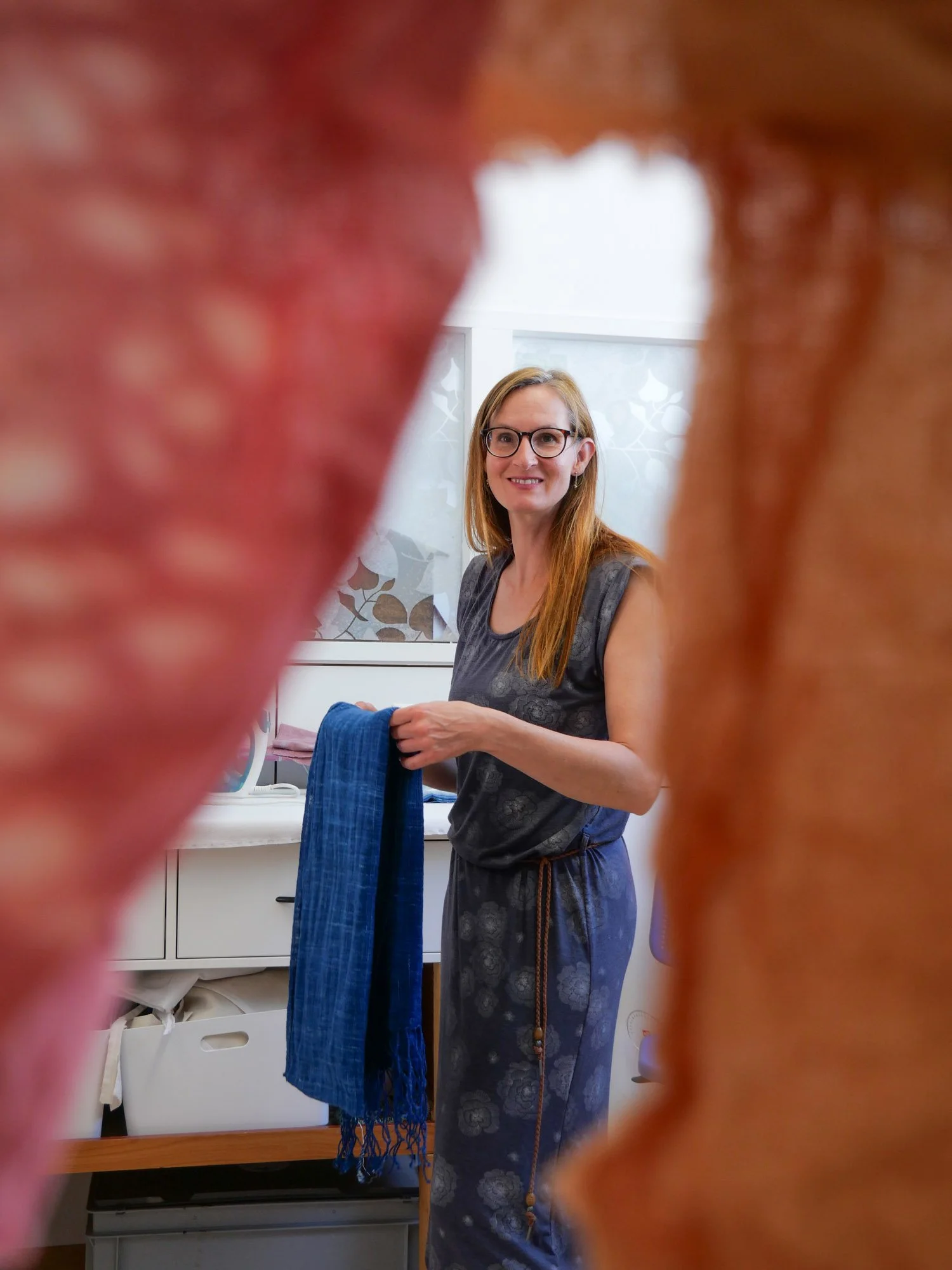 Une femme avec des lunettes et des cheveux roux, souriante, tenant une pièce de tissu bleue, vue à travers un cadre flou autour d'elle.