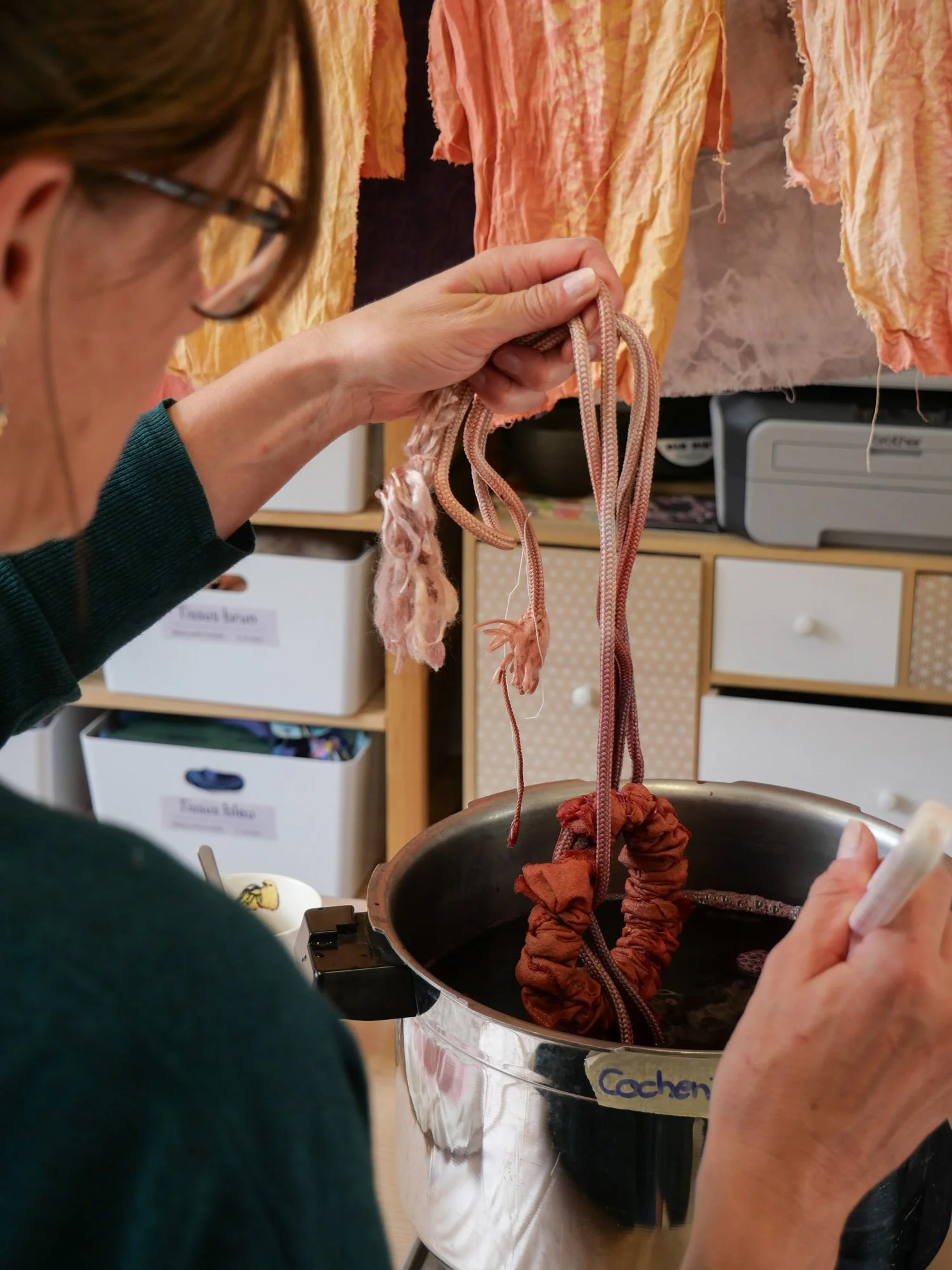 Une personne colorée quelques cordons de tissu dans un bain de teinture, avec des morceaux de tissu suspendus au-dessus.