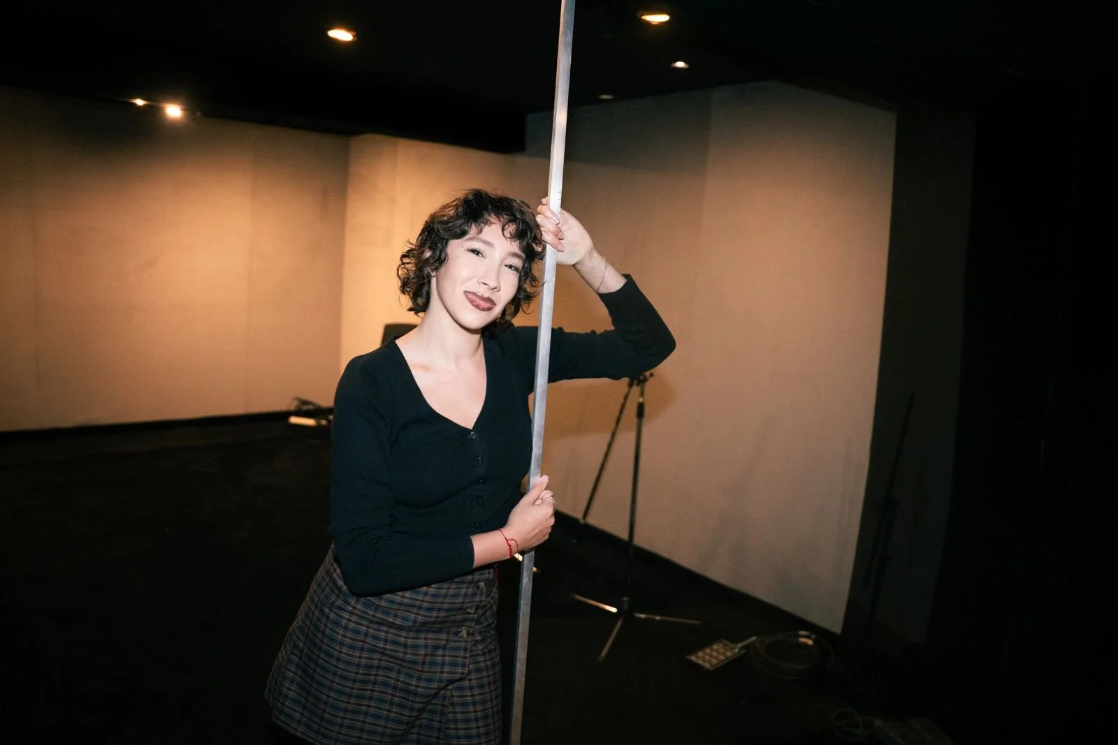 Una mujer con cabello rizado y corto, vestida con blusa negra y falda a cuadros, sostiene una barra en un estudio con fondo oscuro, haciendo una pose confiada.