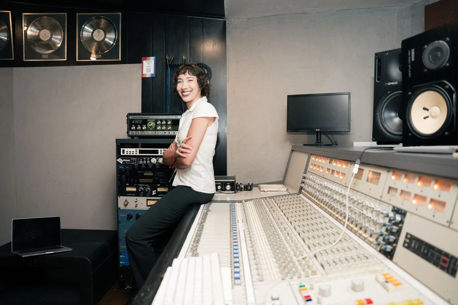Mujer sonriendo en un estudio de grabación rodeada de equipo de audio profesional, incluyendo consolas, monitores y placas de sonido.