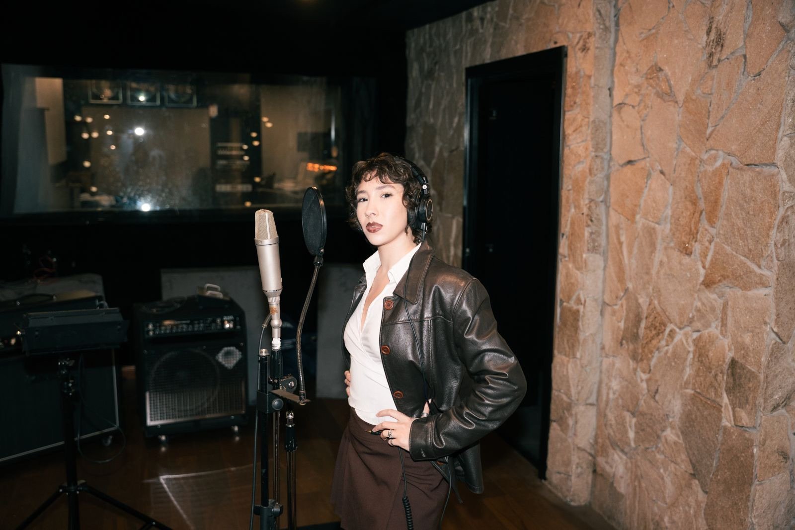 Una mujer en un estudio de grabación, usando audífonos, camisa blanca, chaqueta de cuero y falda marrón, de pie frente a un micrófono, posando con una mano en la cadera.