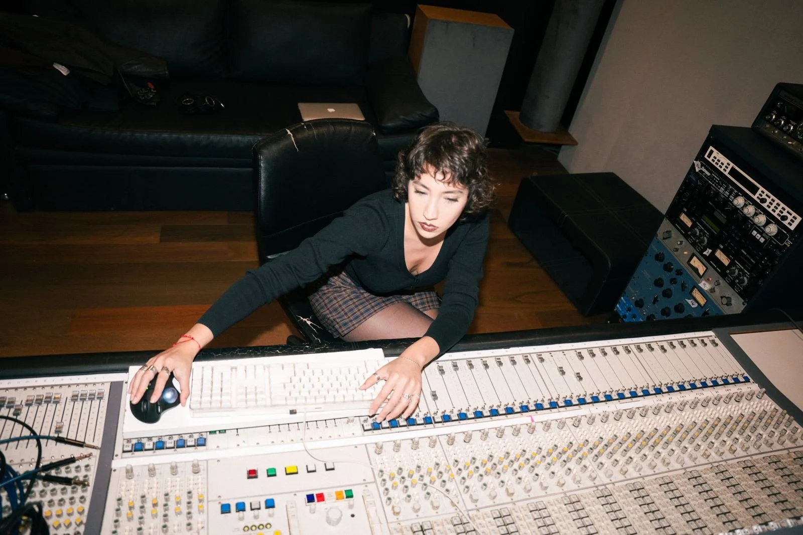 Mujer en estudio de grabación ajustando una consola de audio con un computador en su mano derecha y varios equipos de sonido a su alrededor.