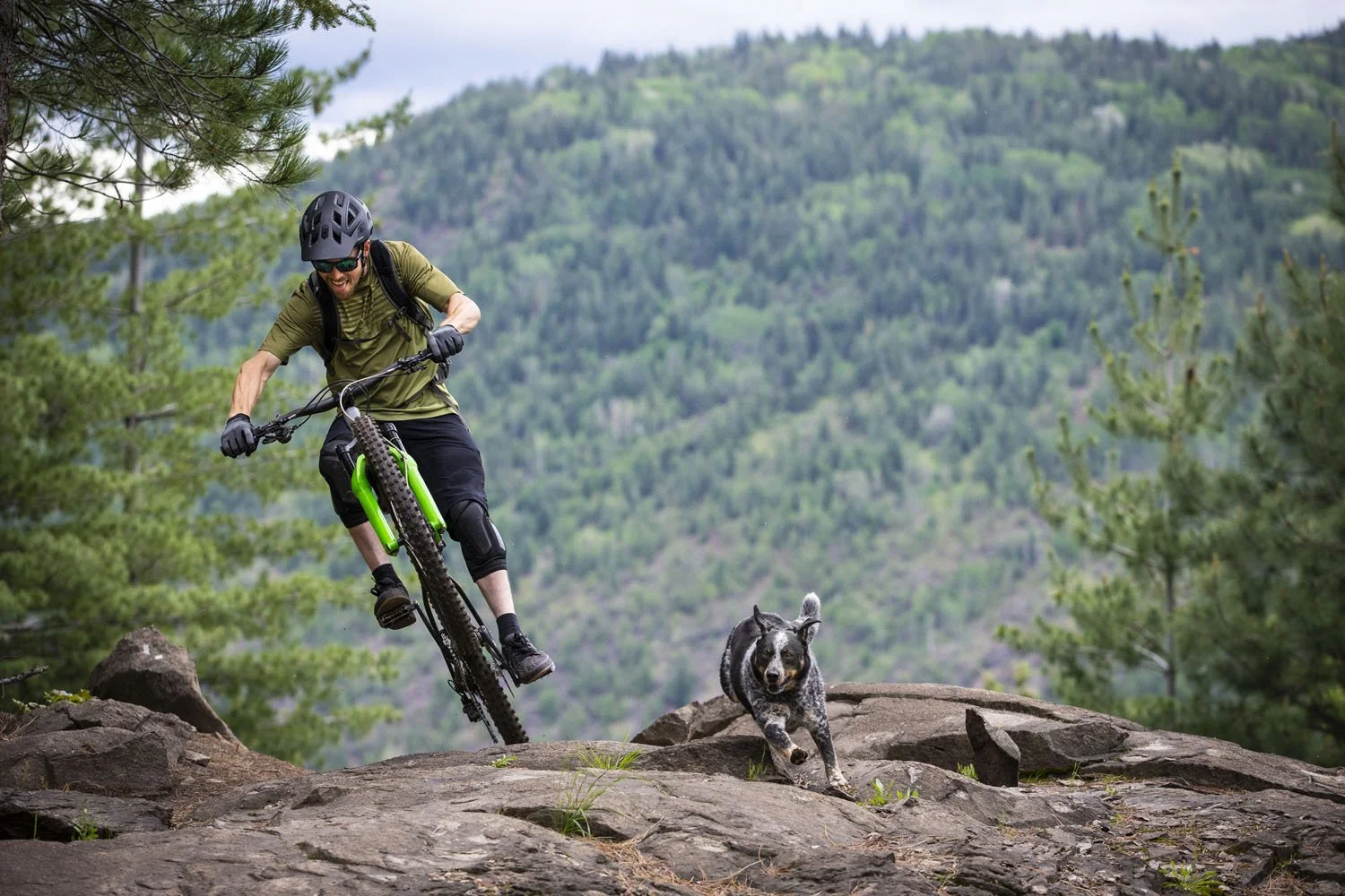 A man riding a mountain bike on rocky terrain with a black and white dog running alongside in a forested mountainous area.