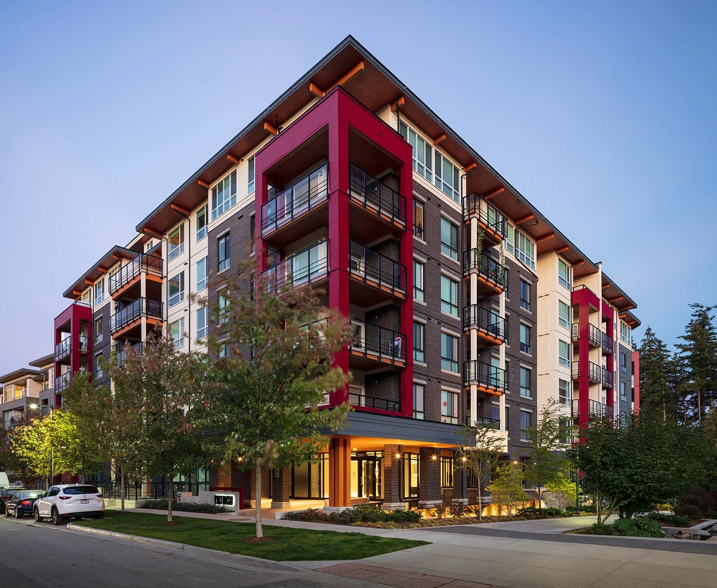 Modern multi-story apartment building with brick and colorful accents, large windows, balconies, landscaped surroundings, and trees in front, taken during dusk.