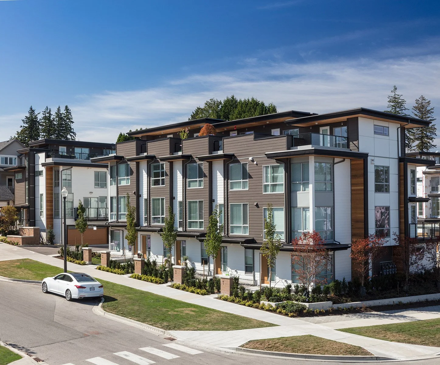 A modern multi-story apartment building with large windows, balconies, and a landscaped sidewalk with trees and a parked white car in front.