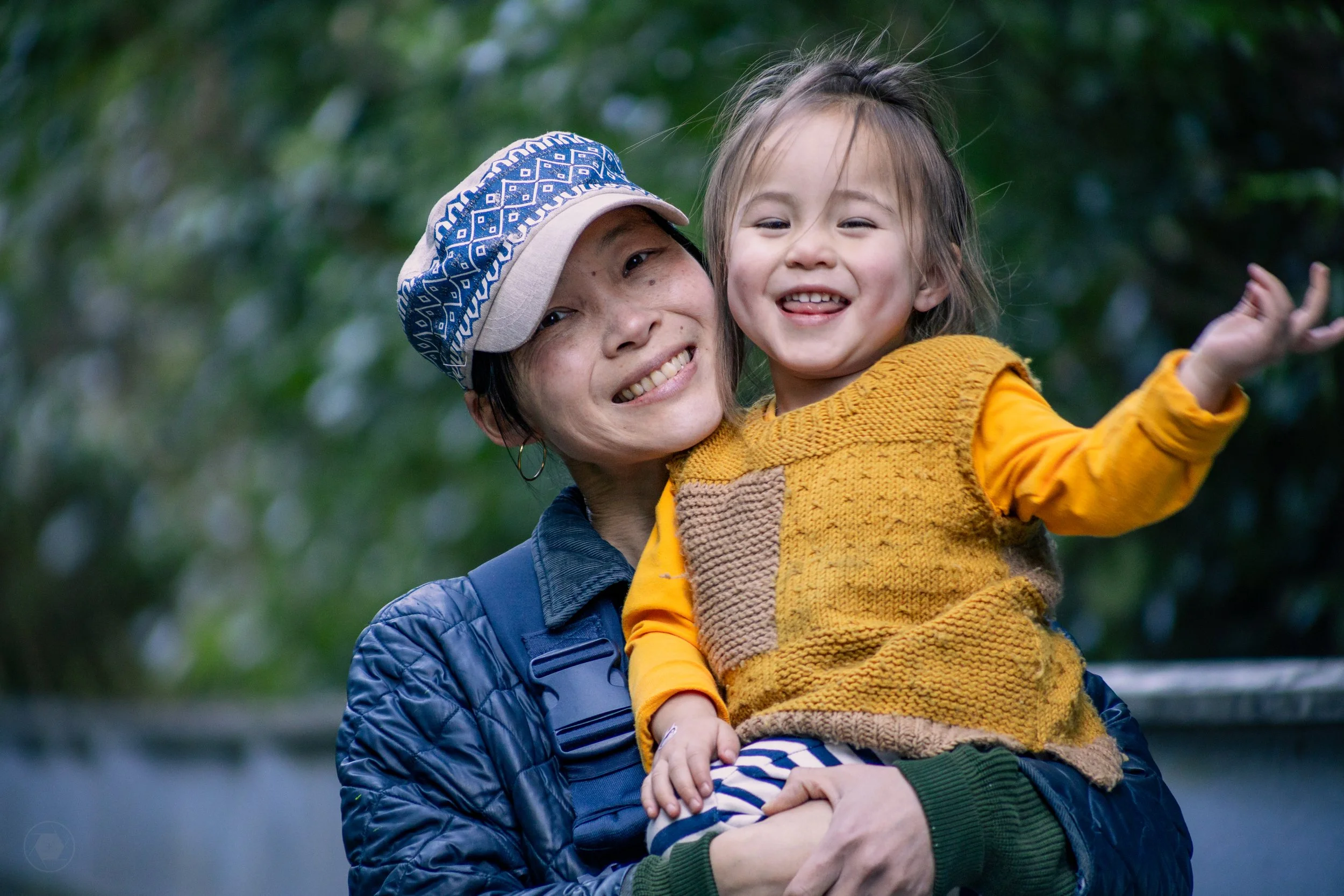 A woman in a blue jacket holds up a child in a yellow cardigan who smiles happily