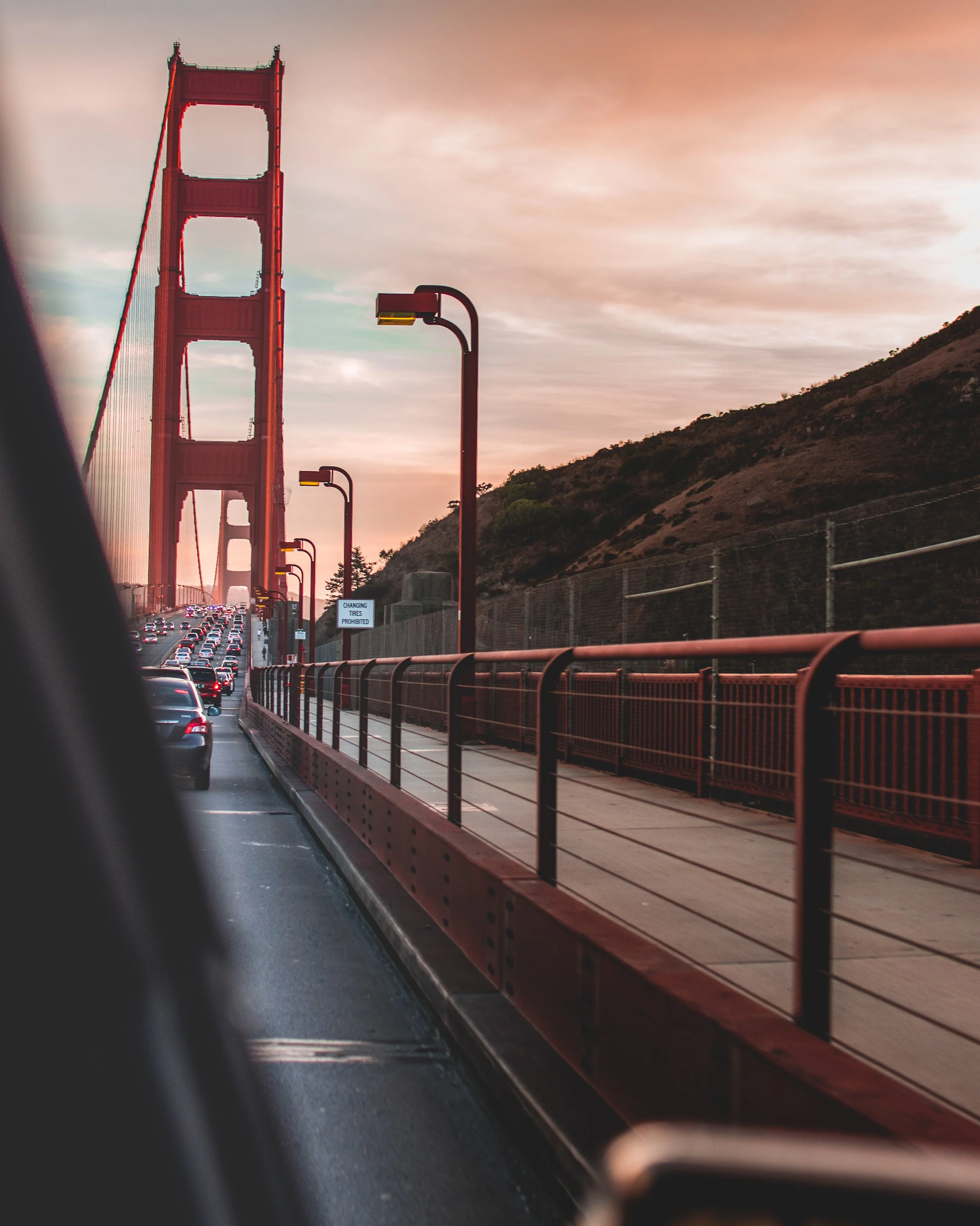 View of the Golden Gate Bridge in San Francisco during sunset, showing cars in traffic on the bridge, with a pedestrian walkway and hillside in the background.