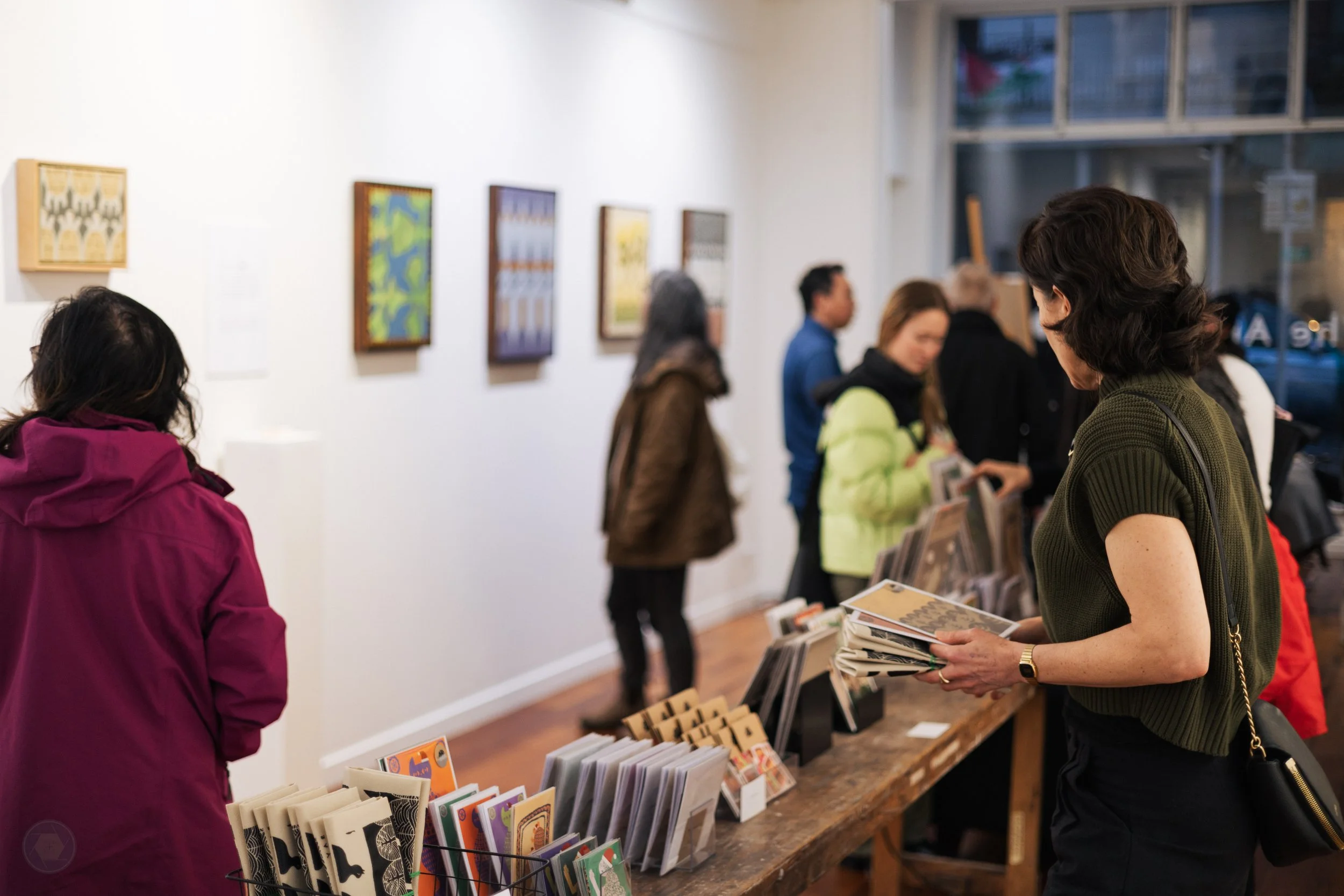 People look around an art gallery and a collection of cards and postcards are on a long counter in the middle of the room