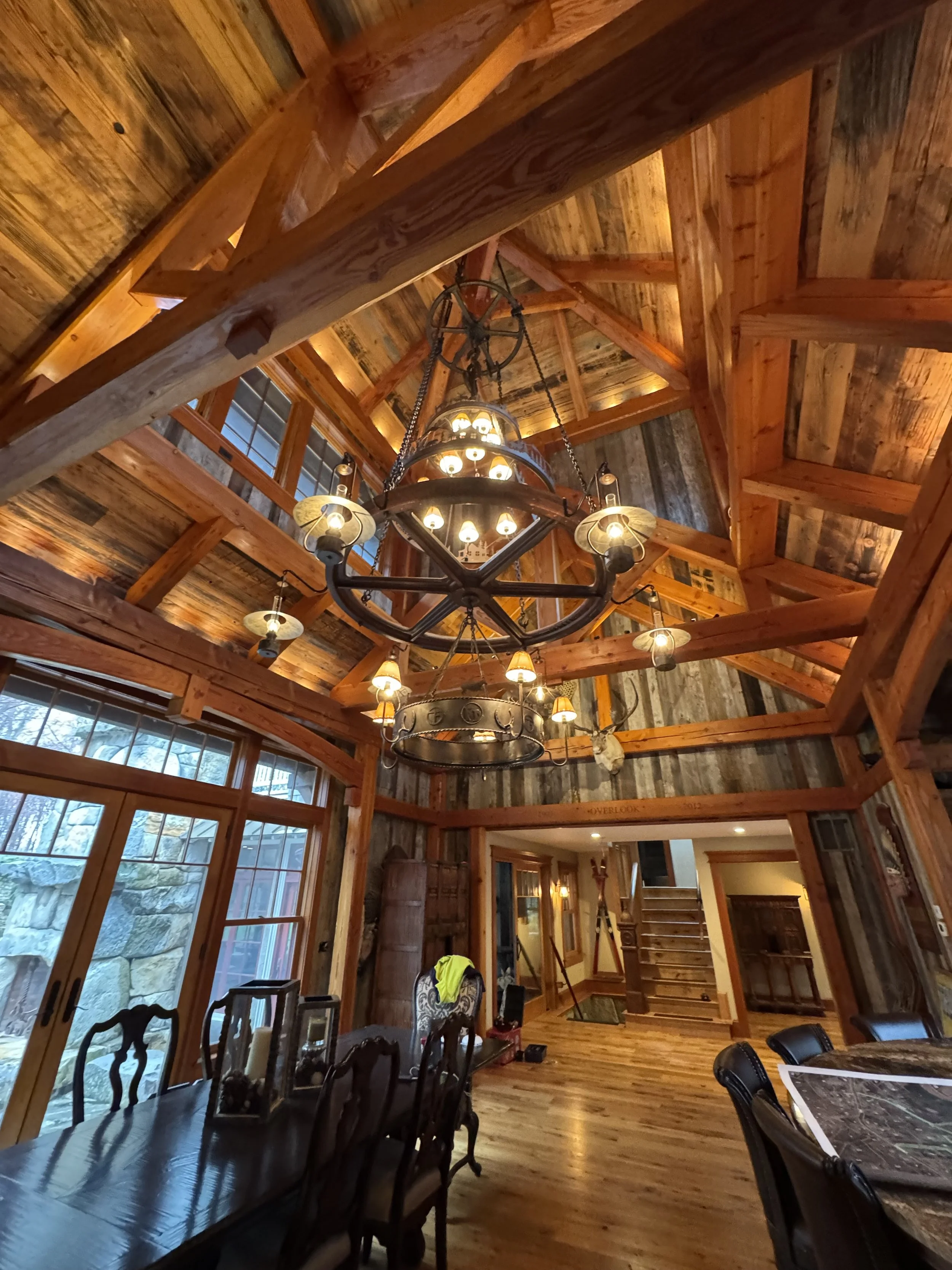 Interior of a rustic wooden cabin with high vaulted ceiling, wooden beams, and a large wrought iron chandelier with multiple small lamps hanging from the ceiling. There is a dining table with chairs and large windows that reveal a stone wall outside.