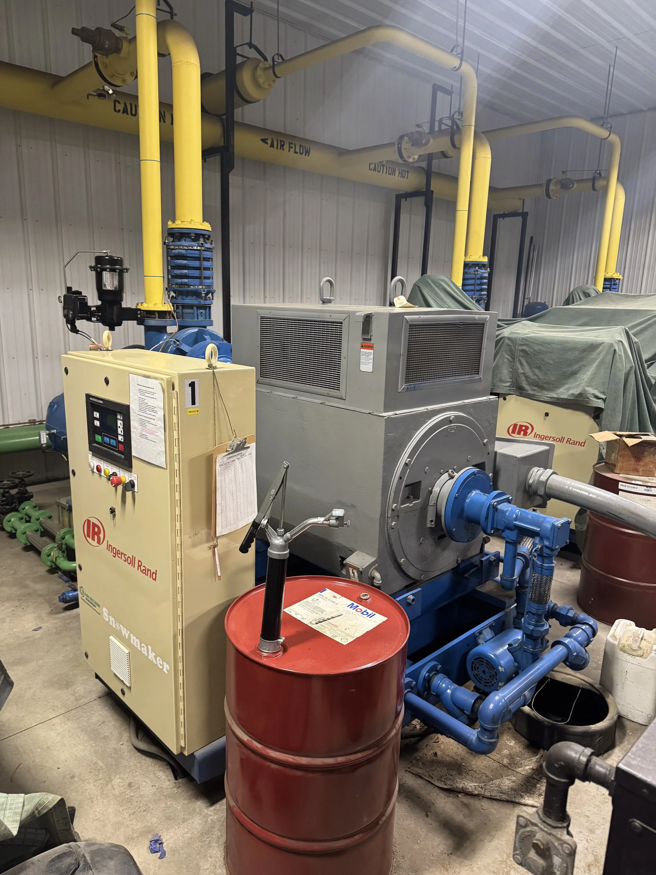Industrial mechanical room with yellow pipes, a large gray machine, and a control panel. A red barrel with a Mobil label is in front of the machinery, surrounded by various pipes and equipment.