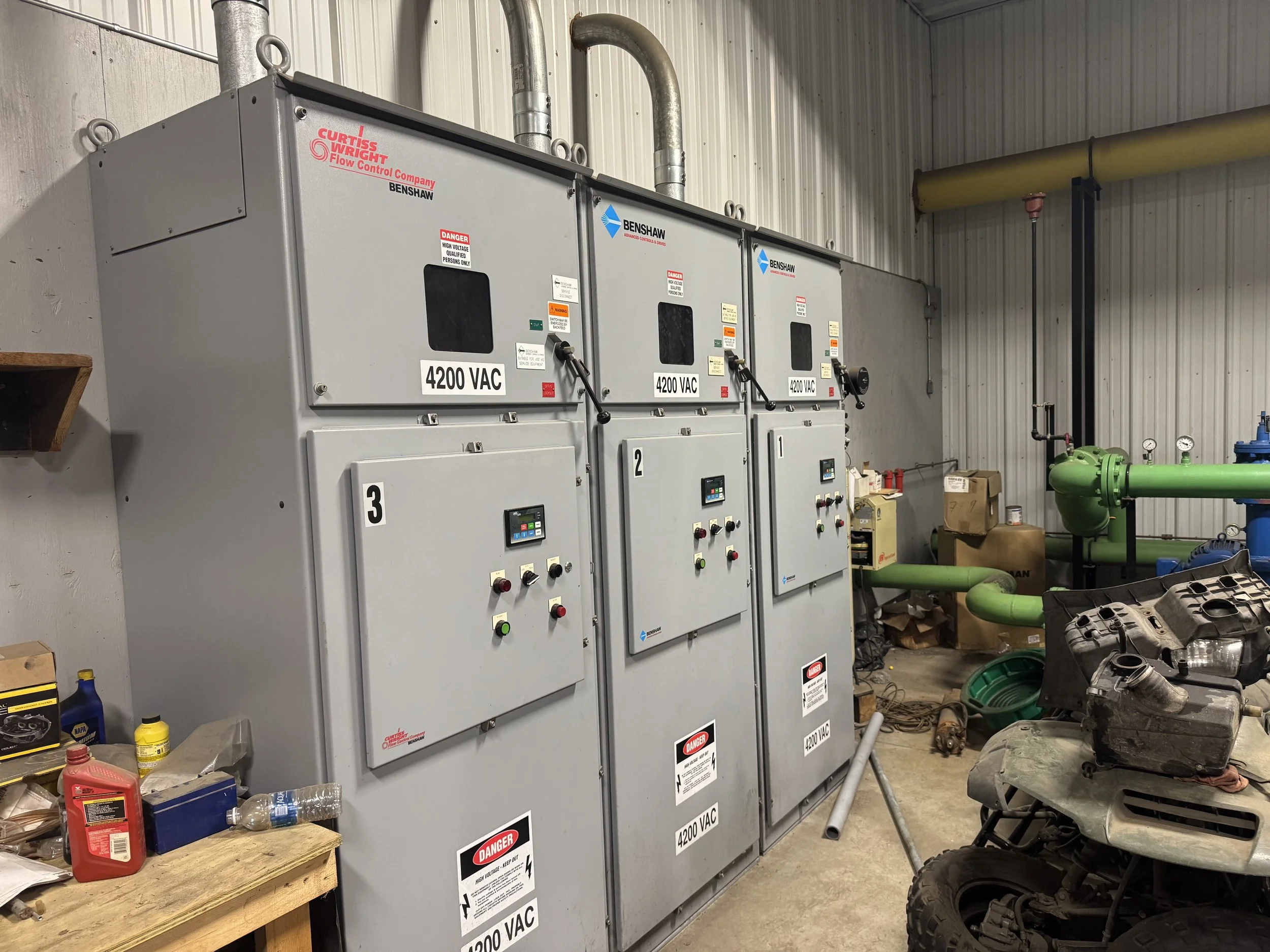 Electrical control panels labeled with voltage ratings and warning signs in an industrial setting, with pipes, tools, and equipment around.