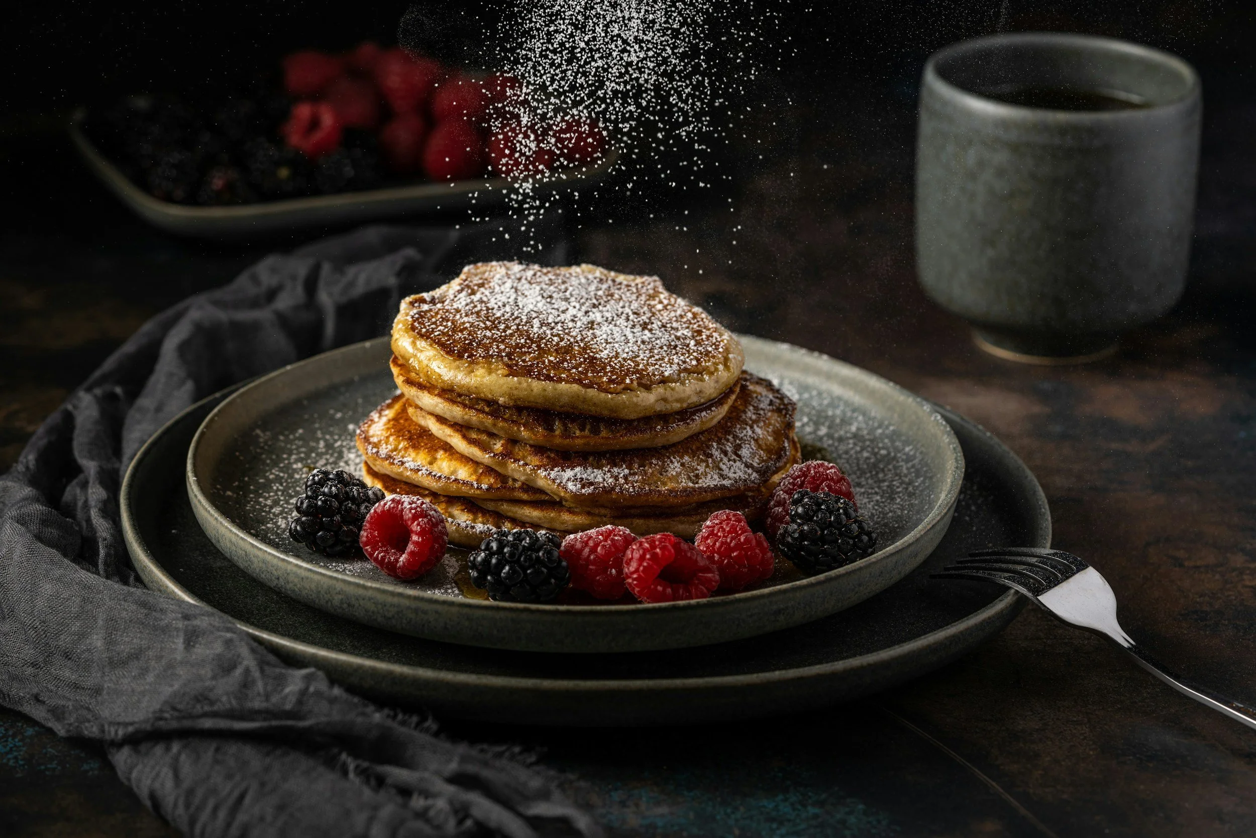 Stacked pancakes dusted with powdered sugar, garnished with blackberries and raspberries, with a cup of coffee in the background.