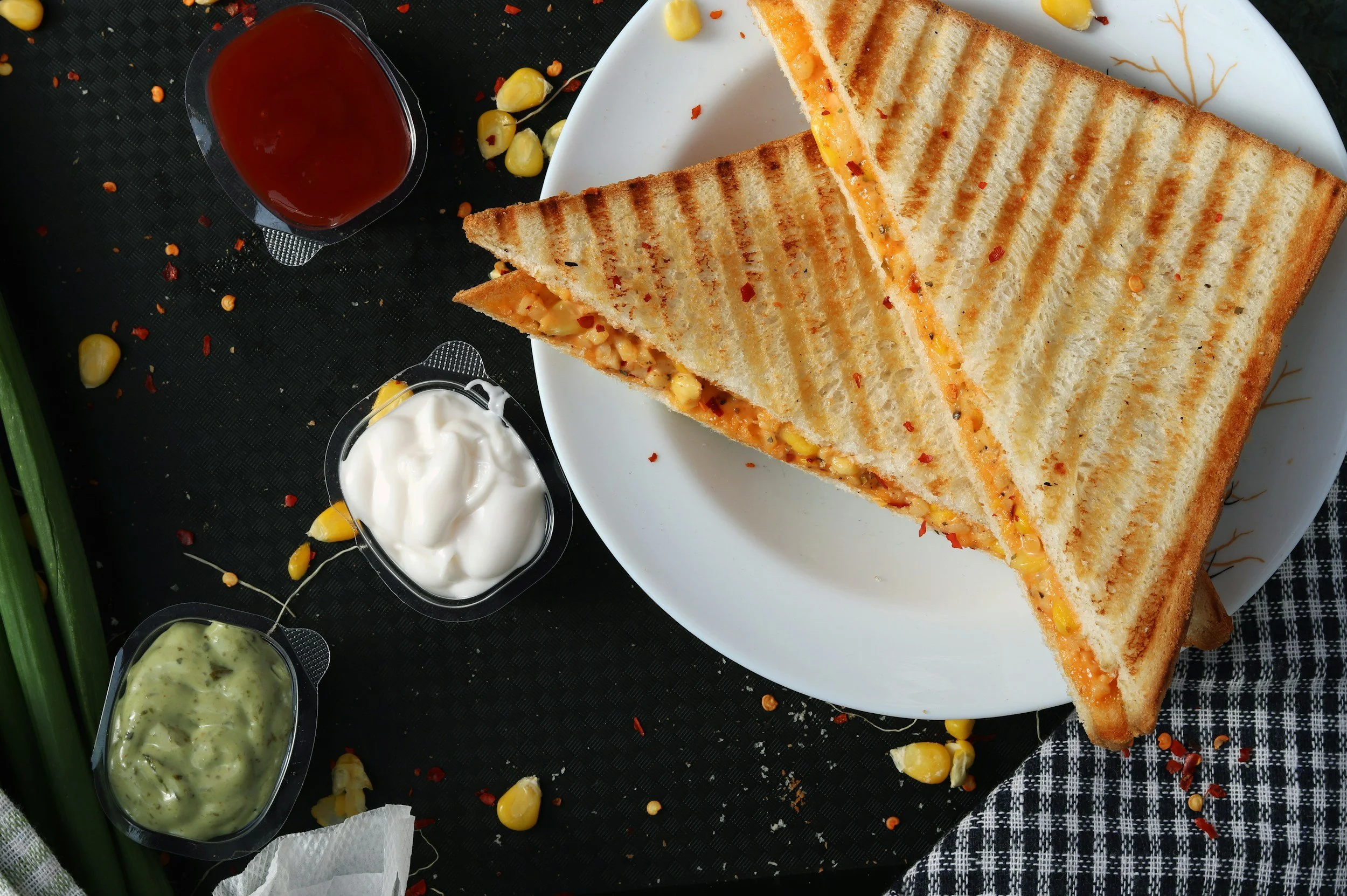 A grilled cheese sandwich cut diagonally on a white plate, with three small containers of condiments (ketchup, sour cream, and green herb sauce) nearby, on a black textured surface with scattered popcorn, chili flakes, and a checkered cloth.