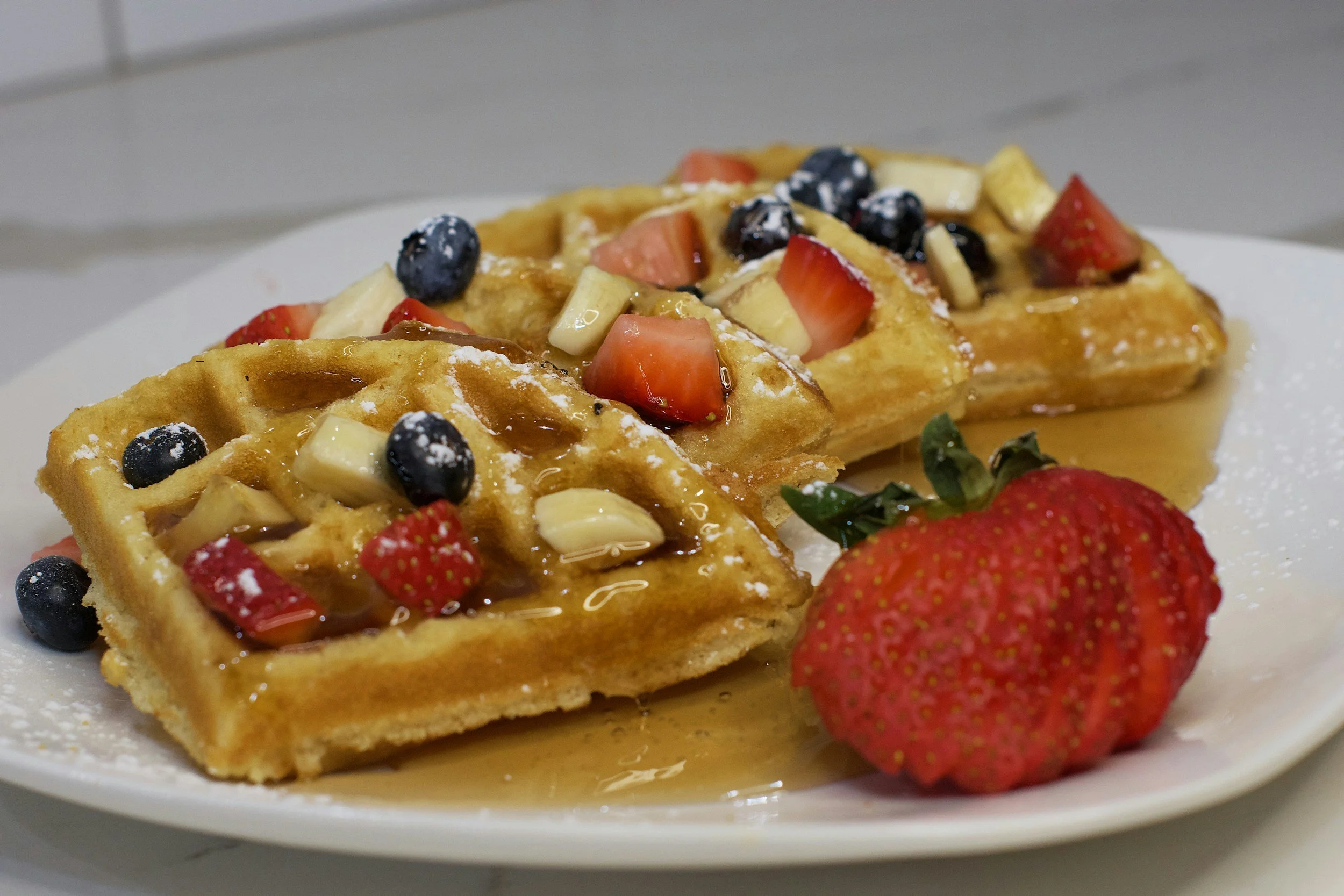 Belgian waffles topped with strawberries, blueberries, white chocolate, and syrup, served with a whole strawberry on a white plate.