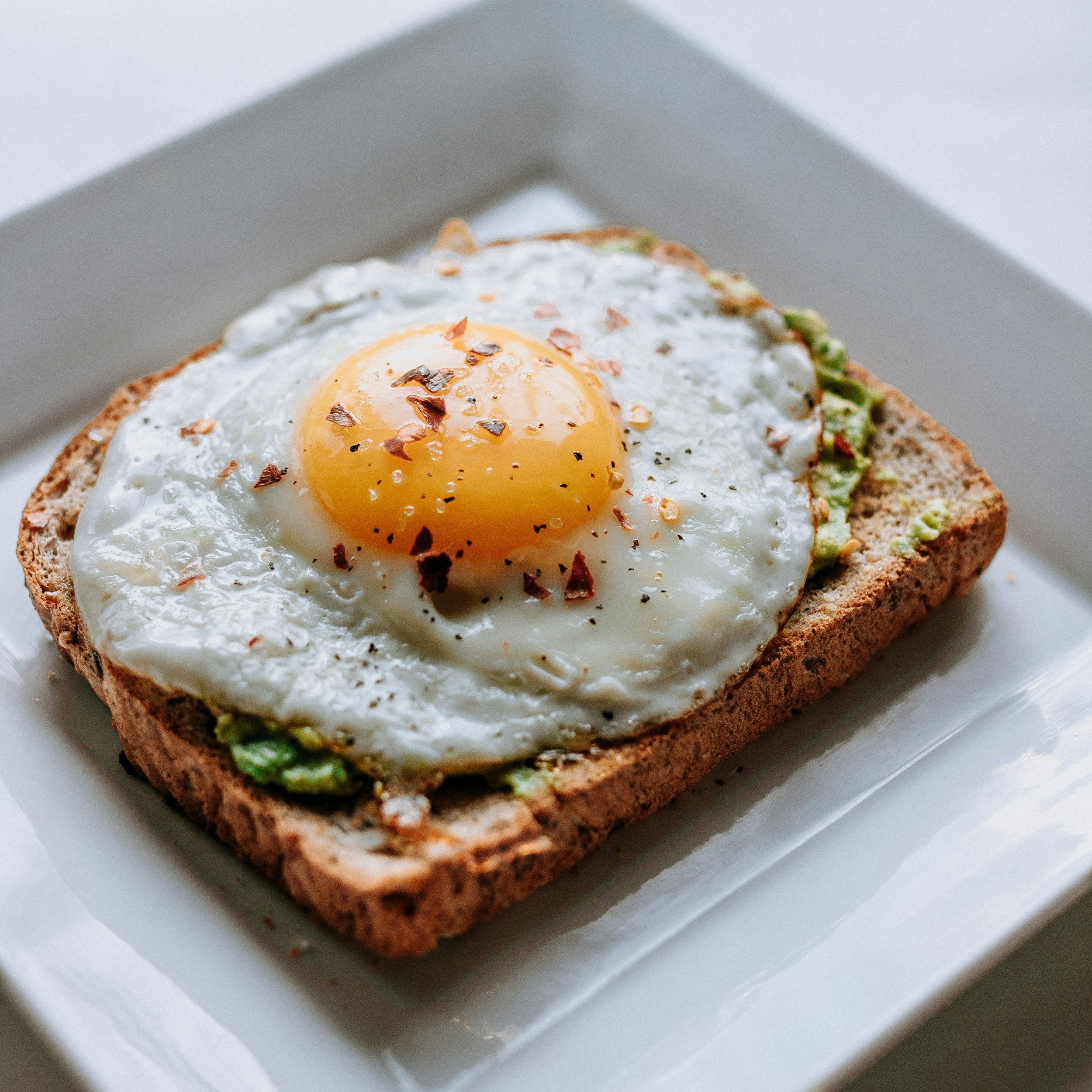 A slice of whole grain toast topped with mashed avocado and a fried egg seasoned with black pepper and red pepper flakes, served on a white plate.