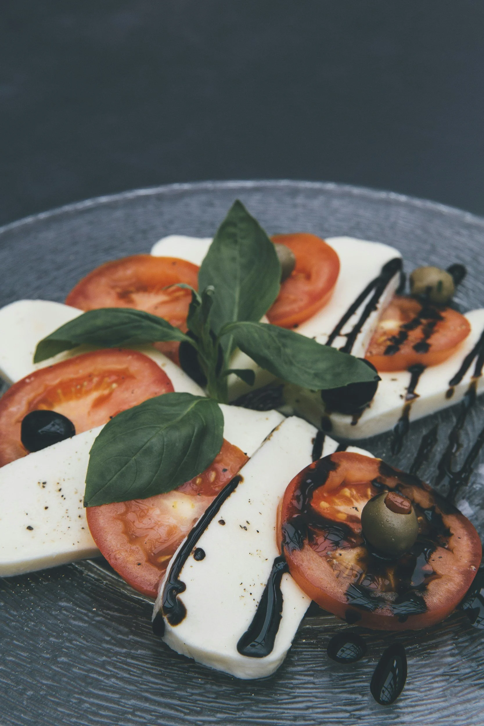 Caprese salad with sliced tomato, mozzarella cheese, basil leaves, black olives, balsamic glaze, and a drizzle of balsamic vinegar on a glass plate.