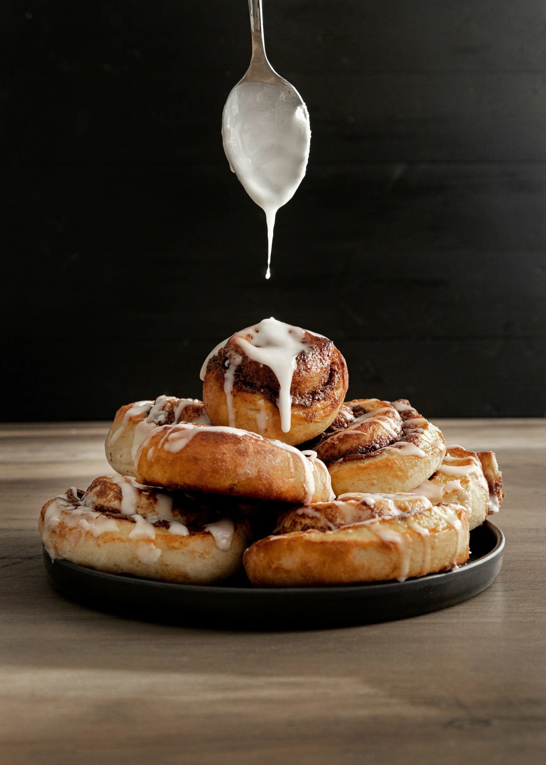 A plate of assorted cinnamon rolls with icing, with a spoon pouring icing over a cinnamon roll above the plate, set on a wooden surface against a black background.