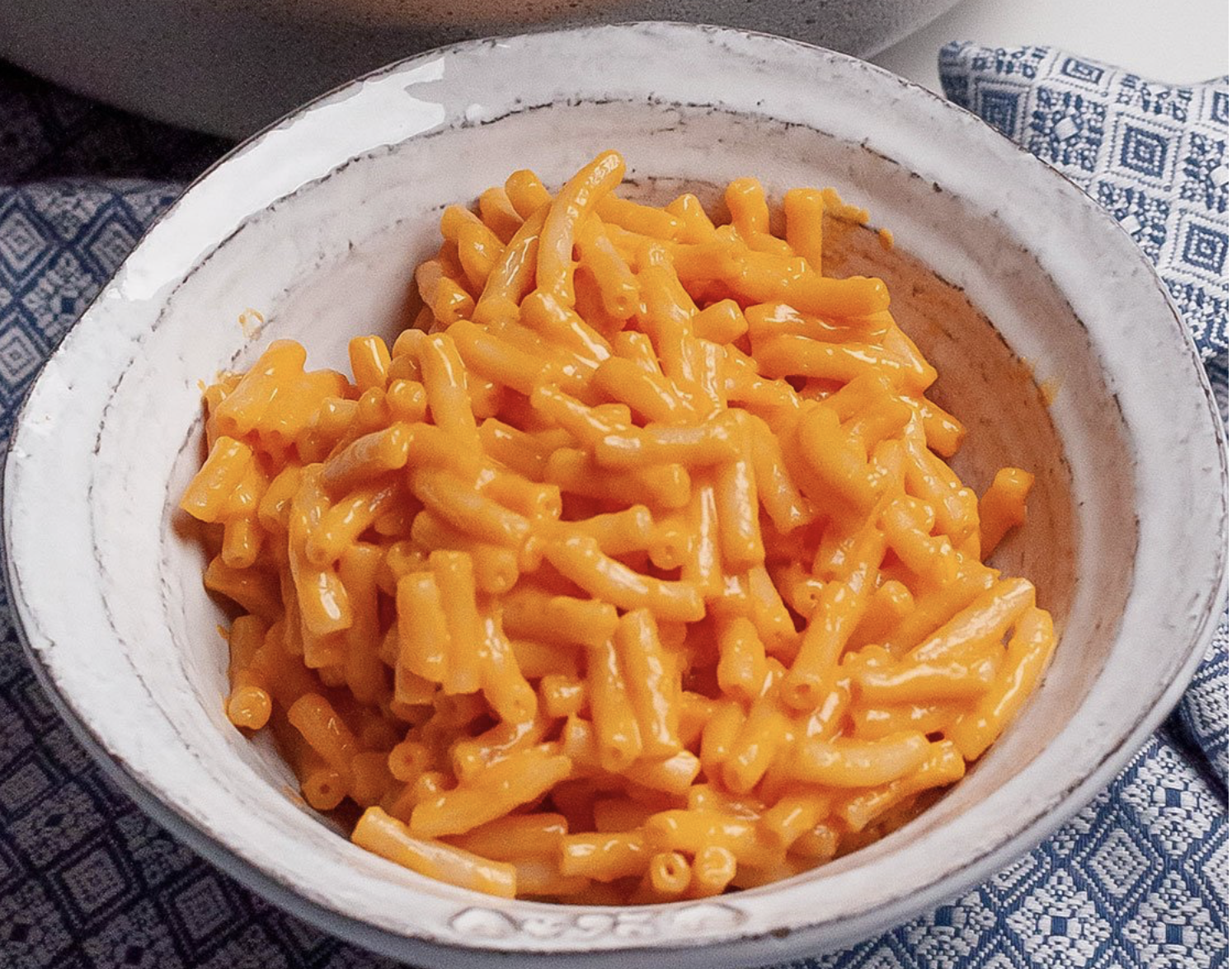 A white ceramic bowl filled with macaroni and cheese on a blue patterned cloth.