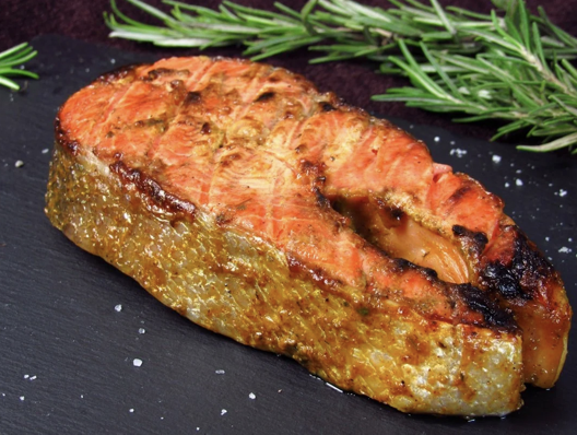 A grilled salmon steak seasoned on a black slate surface with rosemary sprigs and coarse salt.