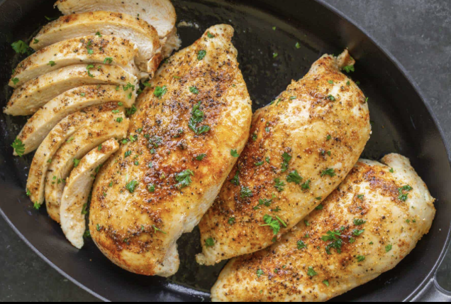 Raw chicken breast on a wooden cutting board surrounded by lemon wedges, garlic cloves, sliced onion, rosemary, and peppercorns.