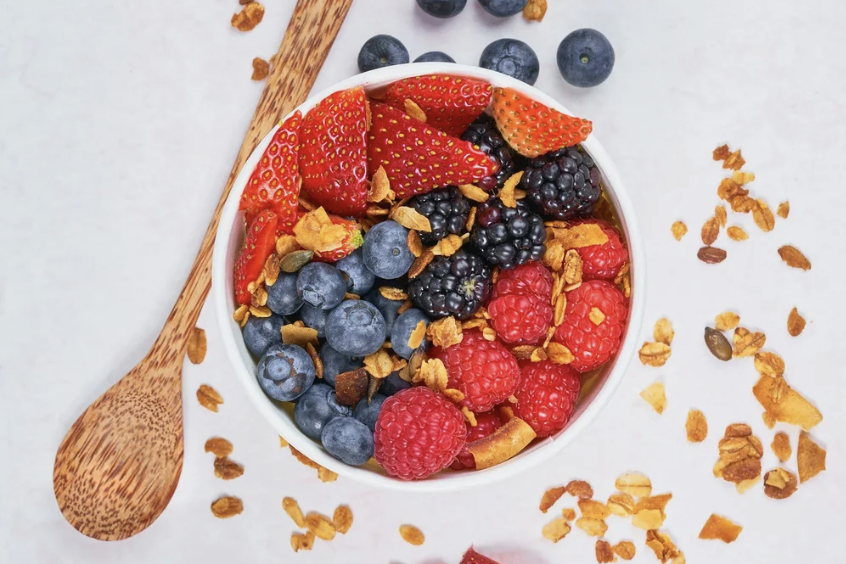 A bowl of mixed fresh berries including strawberries, blueberries, blackberries, and raspberries, topped with granola, with a wooden spoon beside it on a white surface.
