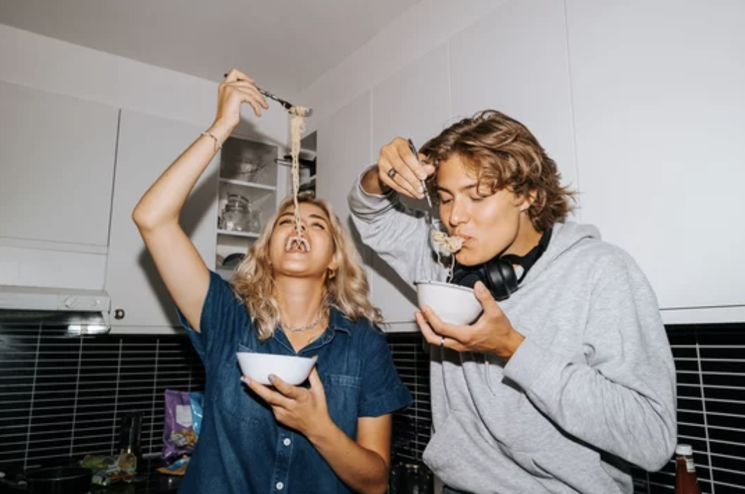 Two young women eating noodles from bowls in a kitchen.