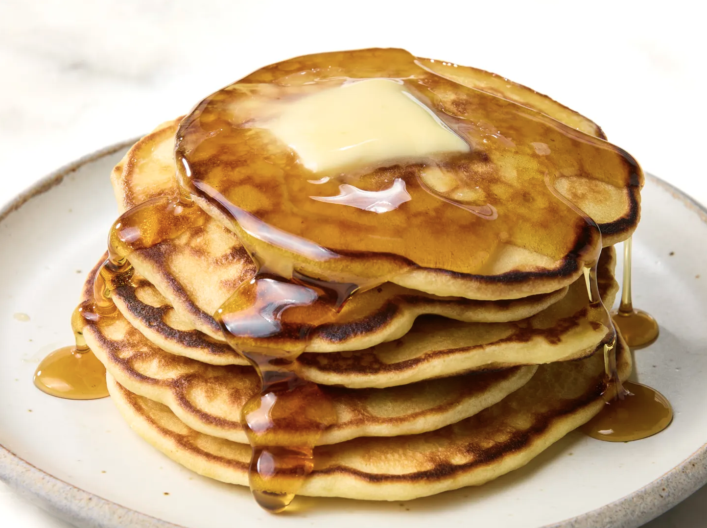 Stack of four pancakes sprinkled with powdered sugar, surrounded by blackberries and raspberries on a plate, with a cup of coffee in the background.