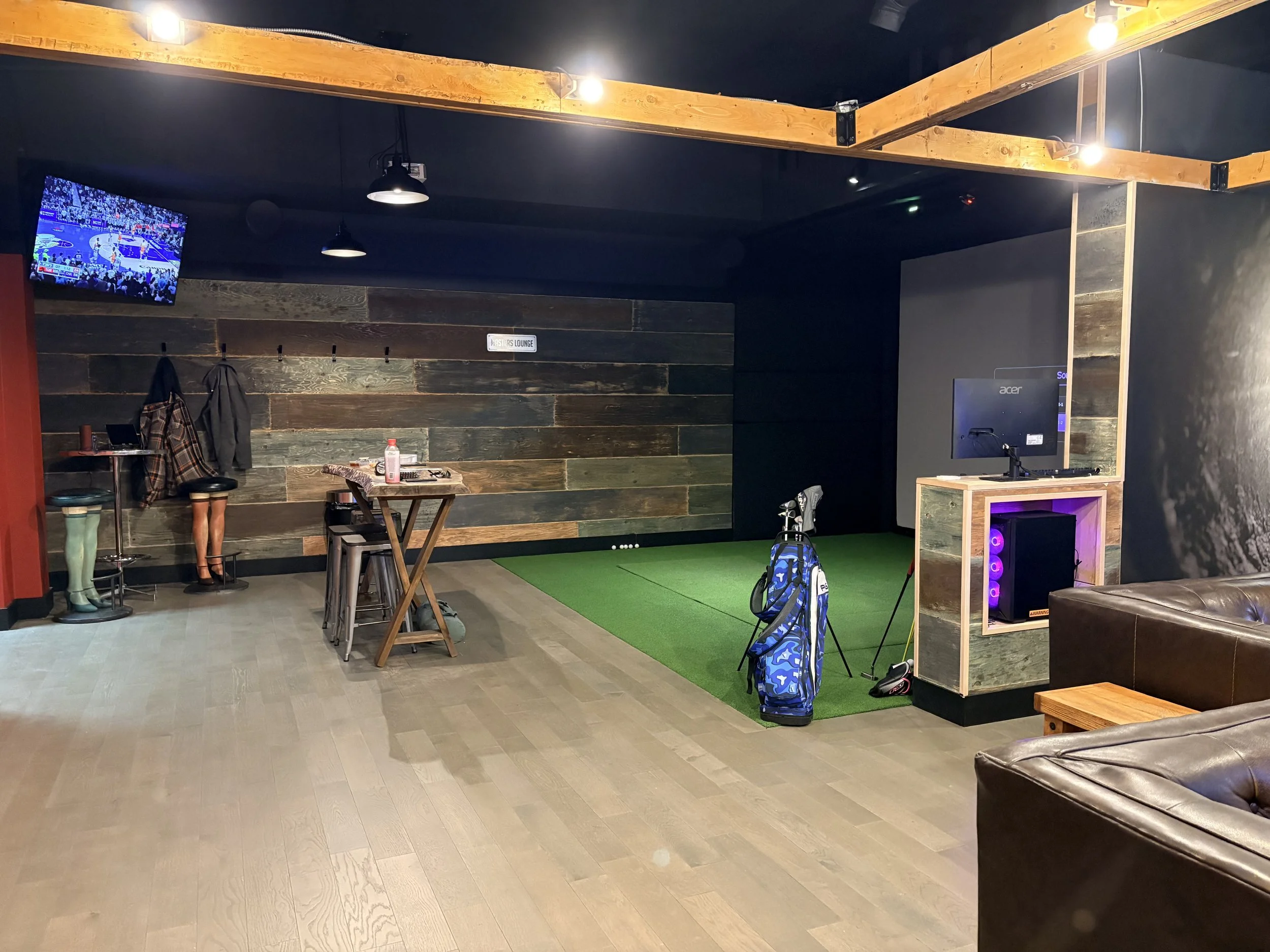 An indoor golf simulation room with a green mat, golf bags, and a computer monitor. A TV is mounted on the wall, and there are leather couches and a small wooden table.