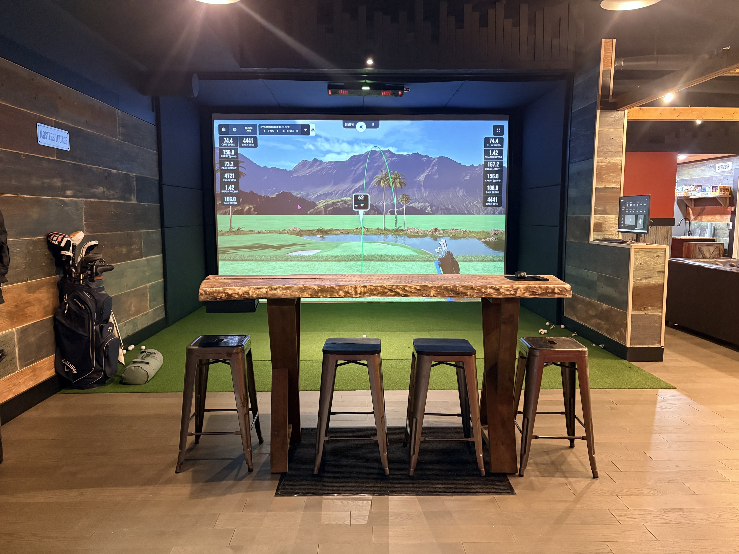 Indoor golf simulator room with a large screen showing a virtual golf course, a wooden table with four stools in front of the screen, golf clubs in a bag on the left, and a few golf balls on the green turf.