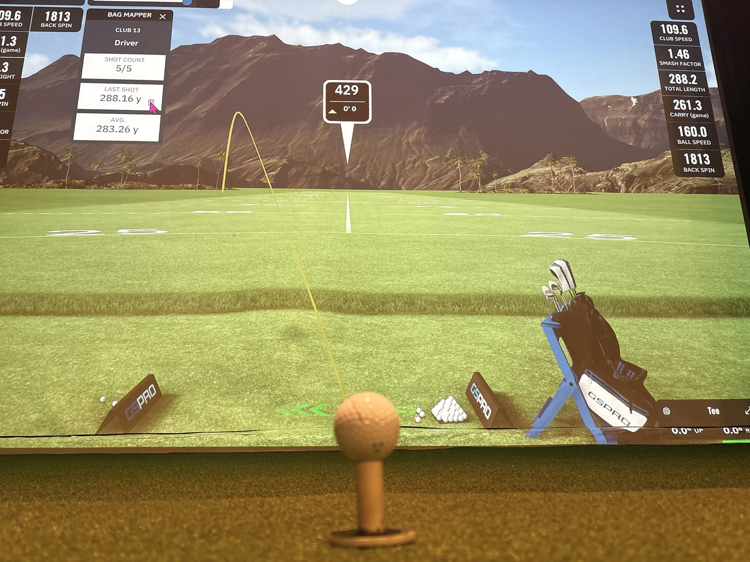 Golf simulation screen showing a virtual golf course with mountains in the background, a golf ball on a tee in the foreground, and various data metrics related to the shot displayed on the screen.