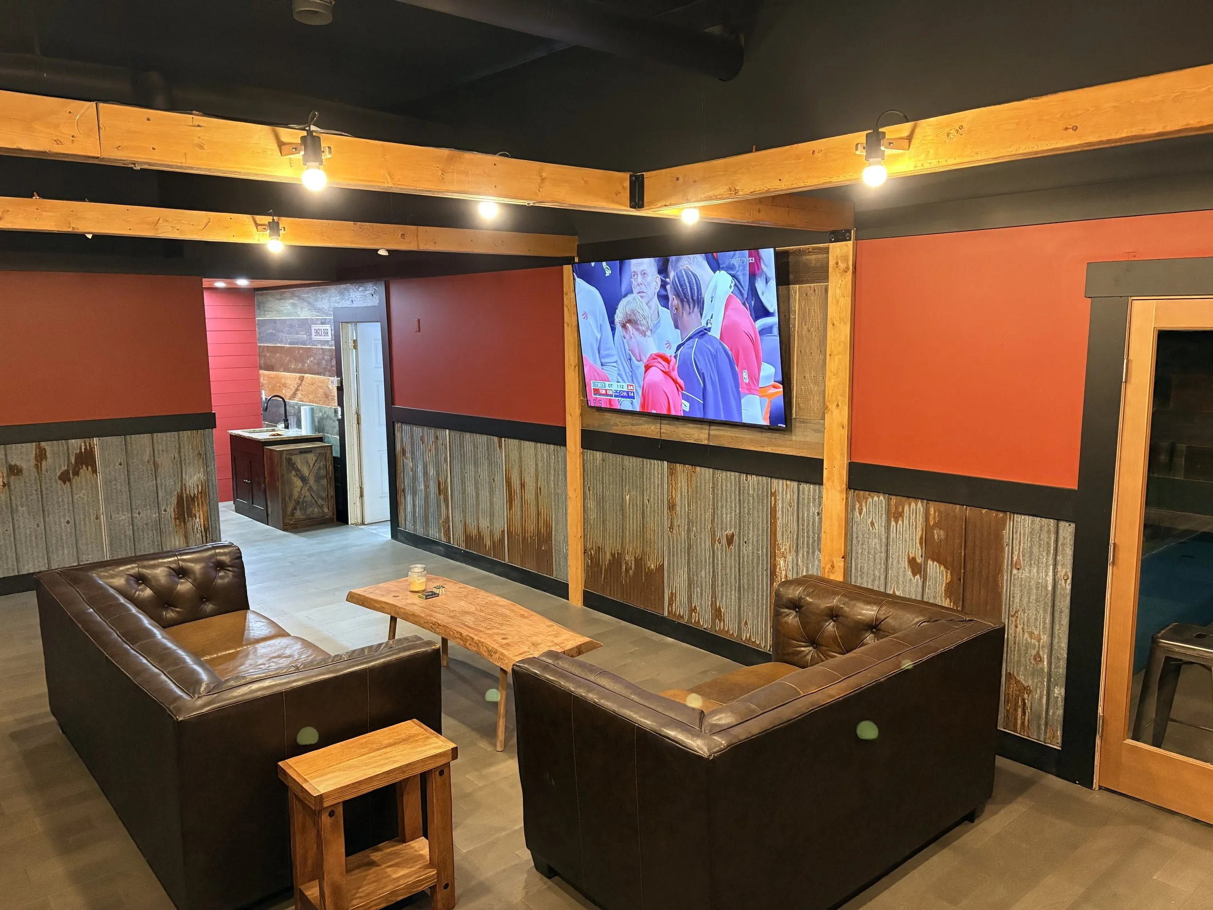 A cozy lounge area with two brown leather couches and a wooden coffee table, a large flat screen TV mounted on a wall with red and rustic paneling, and a small side table nearby. In the background, there is a doorway leading to a room with a kitchenette.