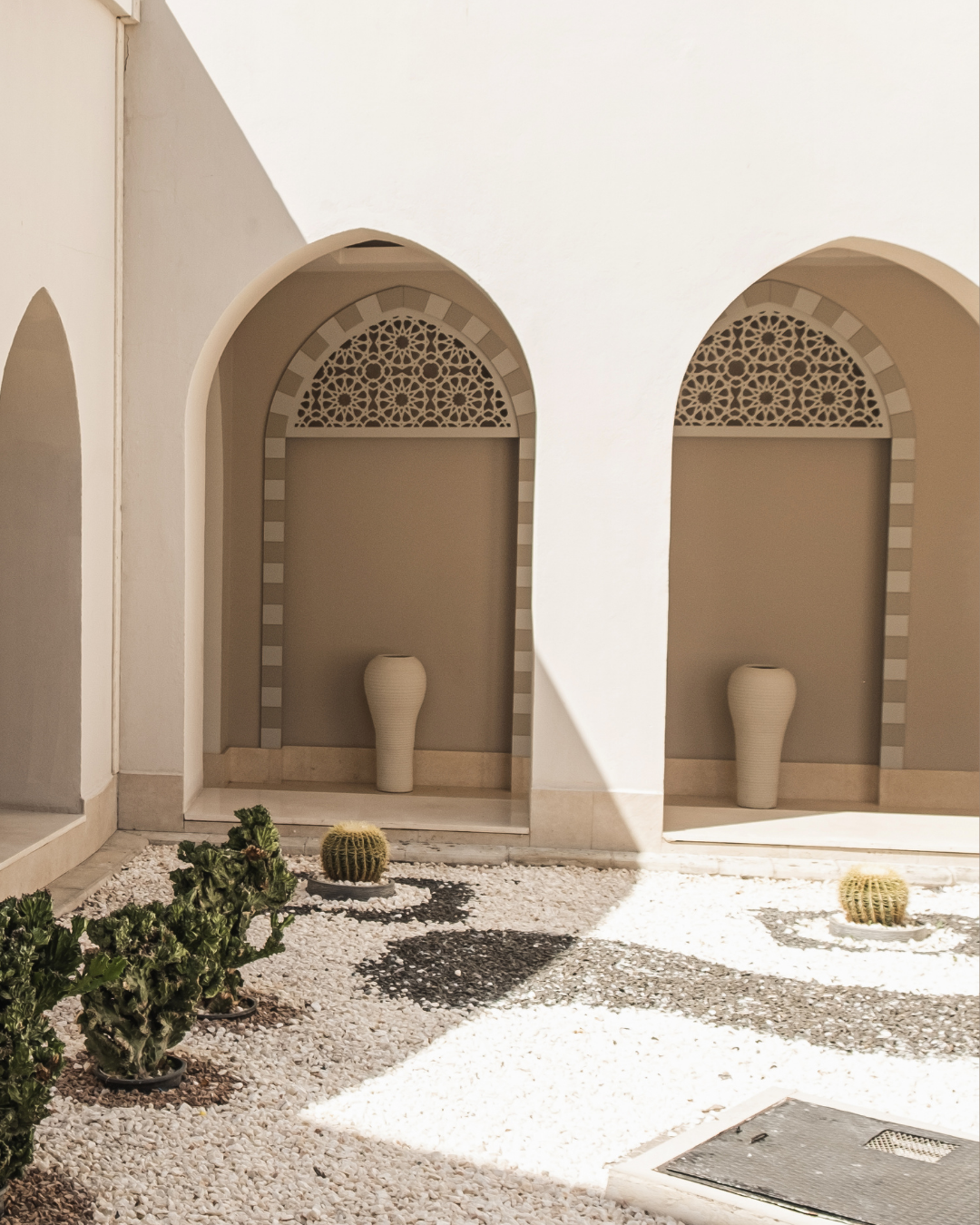 Two arched wall niches with decorative geometric patterns at the top, each containing a ceramic vase. The area includes a small garden with cacti and succulents, and sunlight creates shadows on the white gravel ground.