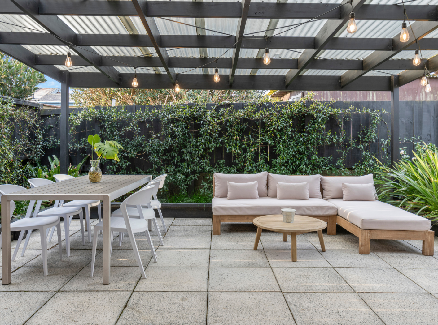 Outdoor patio with a dining table and white chairs to the left, a beige sectional sofa with cushions to the right, a small round wooden coffee table in front, and a black fence with green leafy plants as a backdrop. The patio has a black wooden pergo